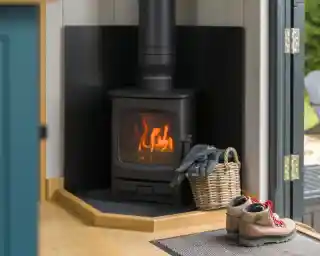 A modern wood stove emits a warm glow, situated in a corner with black backing. Nearby, a woven basket holds blankets, and a pair of rugged boots rests on a mat by the door.