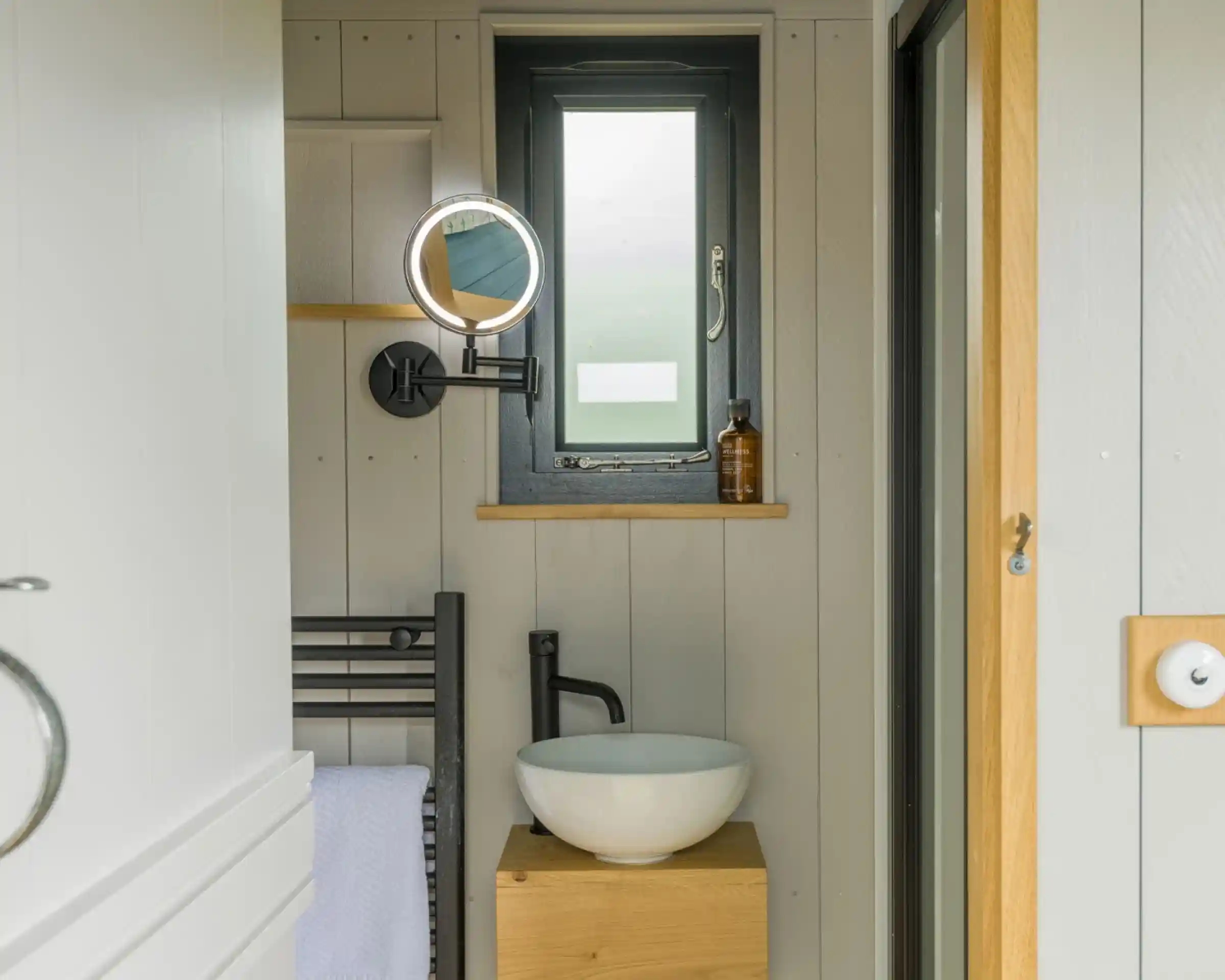 A small bathroom featuring a modern design with a round mirror, a white bowl sink on a wooden stand, and dark wall accents. A window allows natural light in, complemented by a towel rack beside the sink.