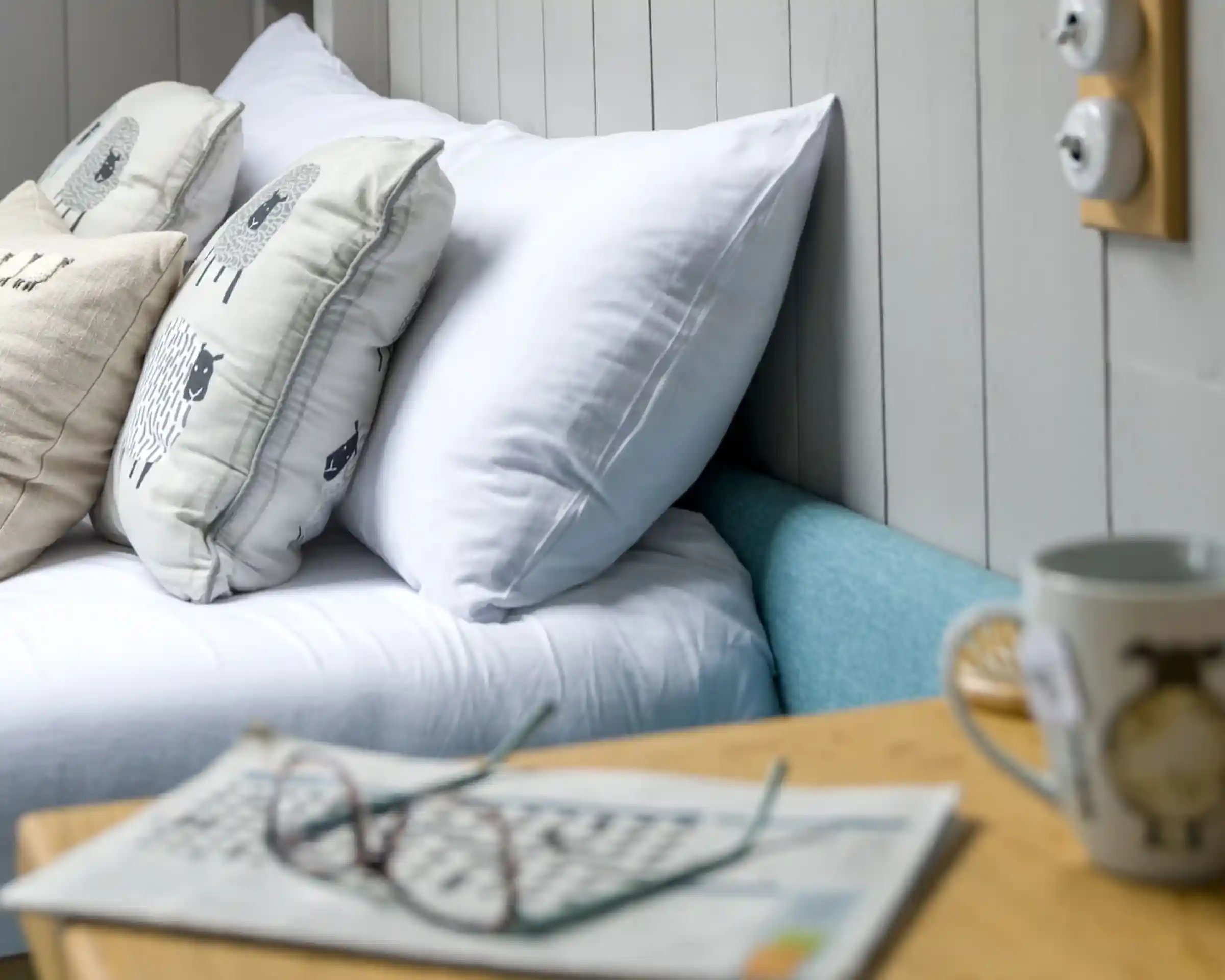 A cozy bedroom scene featuring a bed with white and patterned pillows, a light blue bed frame, a wooden bedside table with a newspaper and glasses, and a mug. Soft light enhances the inviting atmosphere.