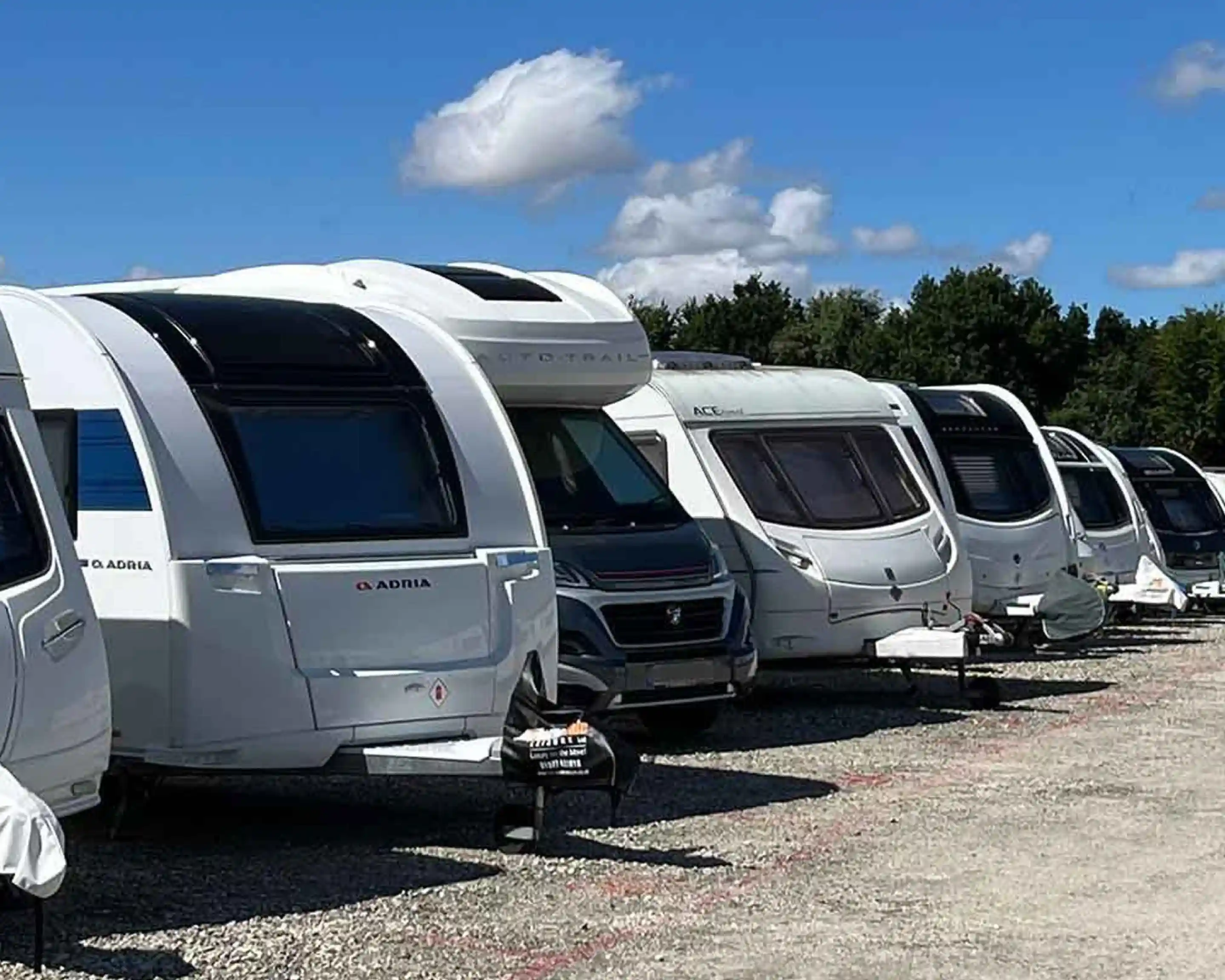 A row of parked caravans lined up on a gravel surface, surrounded by trees and under a blue sky with fluffy clouds.