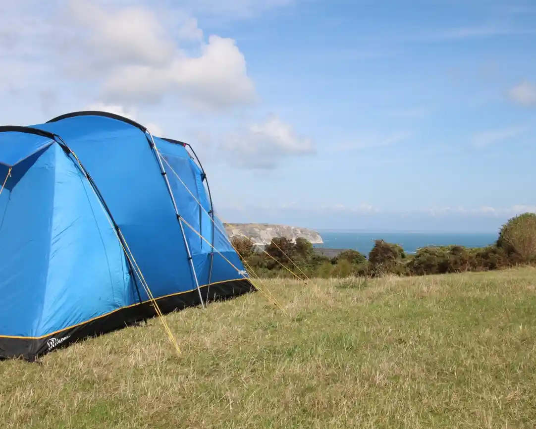 A bright blue tent set up on grassy terrain overlooks a scenic coastline under a partly cloudy sky. The sea is visible in the distance, along with rugged cliffs.