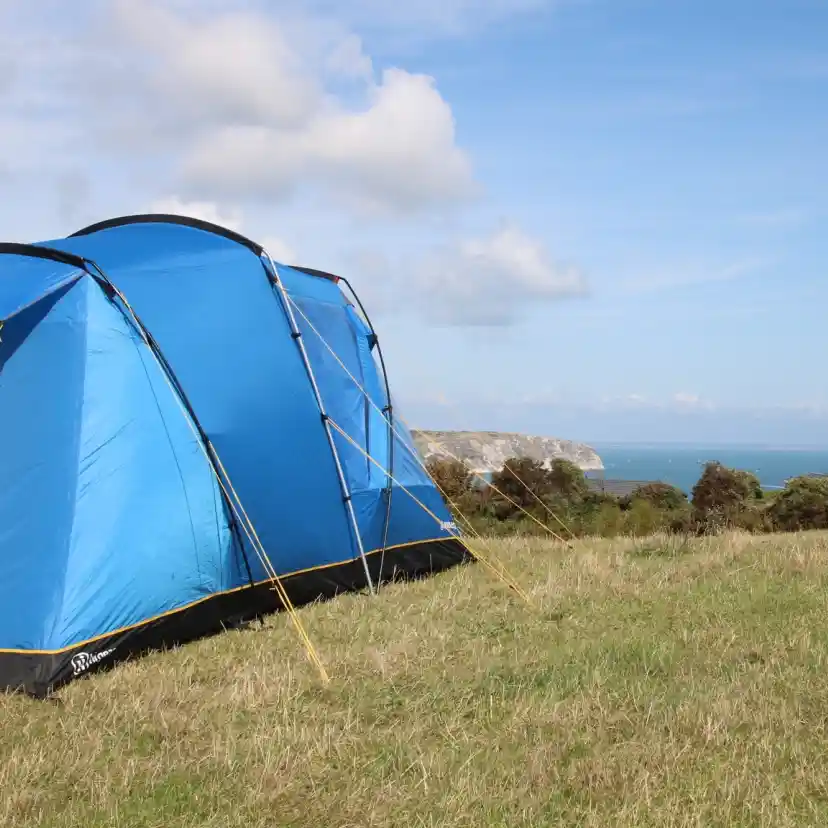 A bright blue tent set up on grassy terrain overlooks a scenic coastline under a partly cloudy sky. The sea is visible in the distance, along with rugged cliffs.
