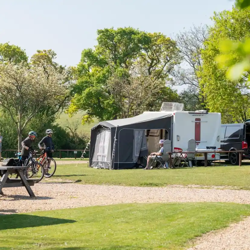A campsite featuring several caravans and a large awning. Two cyclists stop near a picnic table, while others relax in chairs. Lush green trees and a clear blue sky complete the tranquil scene.