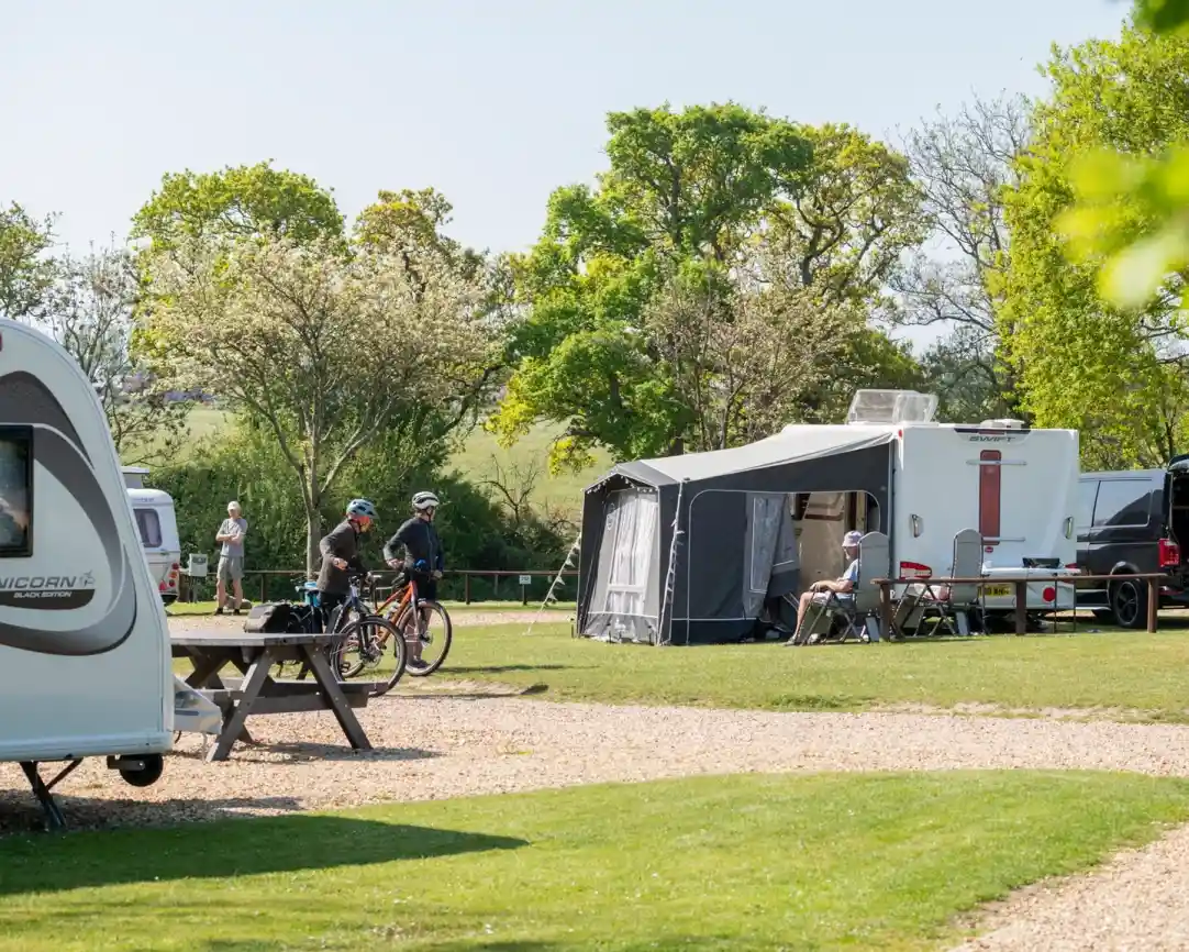 A campsite featuring several caravans and a large awning. Two cyclists stop near a picnic table, while others relax in chairs. Lush green trees and a clear blue sky complete the tranquil scene.
