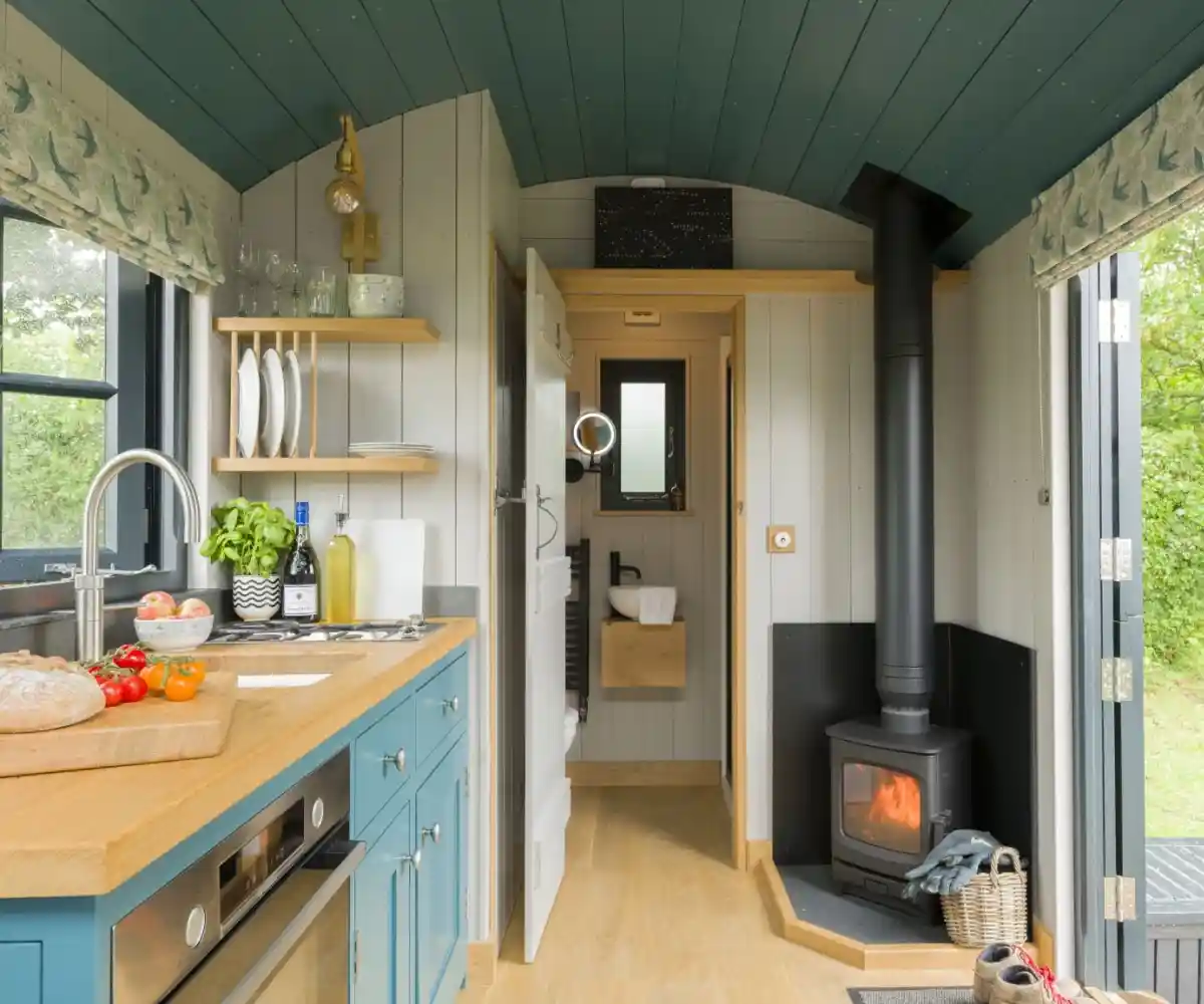 A couple stands on a balcony with a modern home in the background. The man and woman are smiling, dressed casually, with green grass and a landscaped area surrounding the house. The sky is clear and blue.