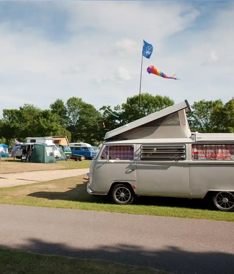 Aerial view of a campsite with grassy areas, trees, and several parked caravans. In the background, the coastline is visible along with a distant town and fields. Clear blue sky enhances the scenic landscape.