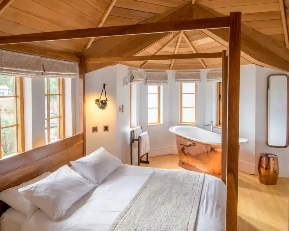 A cozy interior of a well-lit room featuring a wooden four-poster bed with white bedding, a round copper bathtub, and large windows. Warm wood accents create a relaxed atmosphere.