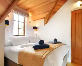 A cozy bedroom with a wooden ceiling, featuring a large bed adorned with a beige quilt and neatly arranged towels. Soft lighting from wall sconces enhances the warm atmosphere. A window lets in natural light, and a wooden door is visible in the background.