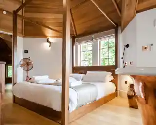 A cozy bedroom featuring a wooden four-poster bed with crisp white linens, a small fan, and large windows allowing natural light. The room has a warm, inviting atmosphere with wooden beams and a minimalist design.