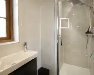 A modern bathroom with a shower, sleek grey tiles, a white vanity sink, and a wooden window frame allowing natural light.