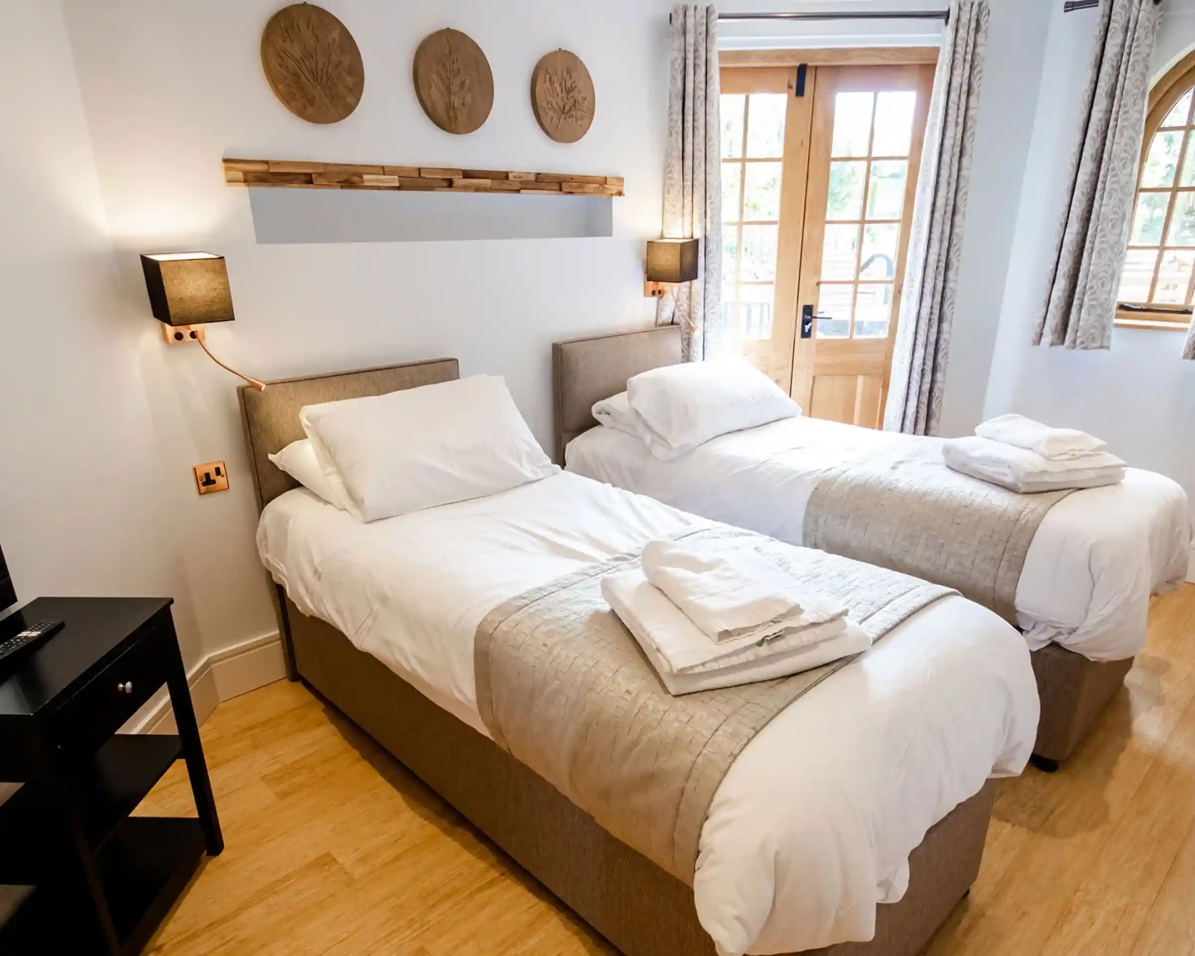 A cozy hotel room featuring two twin beds with light bedding, neatly arranged towels on each bed, wooden accents, and a door leading to an outdoor area. Natural light filters in through the windows, enhancing the warm, inviting atmosphere.