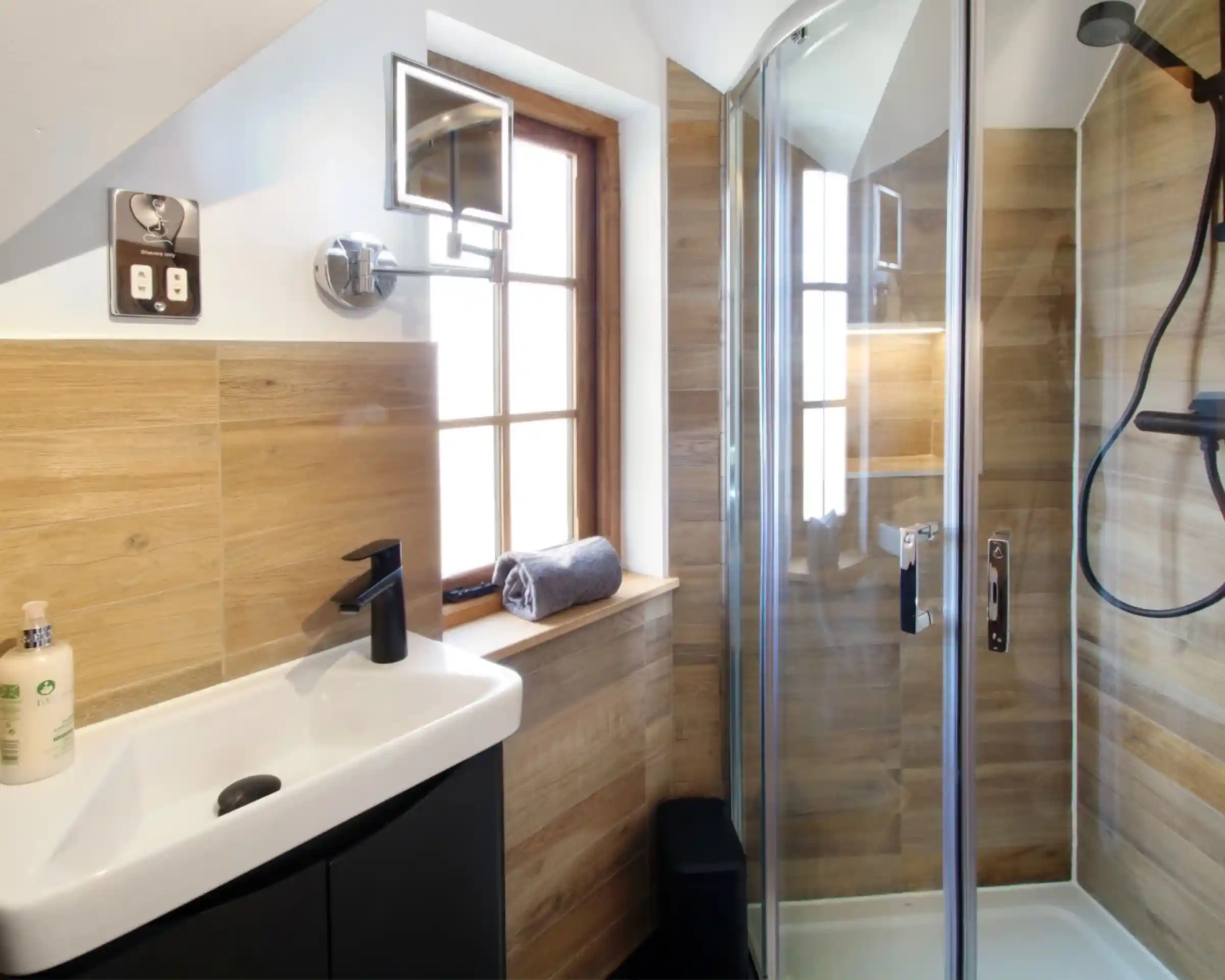 A modern bathroom featuring wooden wall panels, a sleek black sink with a faucet, and a glass shower enclosure. Natural light filters through a window above the sink, while a rolled towel and a black storage container are visible.