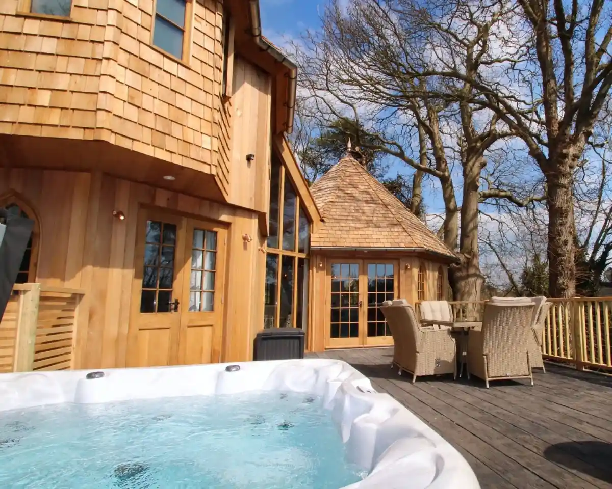 A wooden deck features a hot tub in the foreground, with a charming, rounded wooden building in the background surrounded by trees. Sunlight illuminates the scene under a clear blue sky.