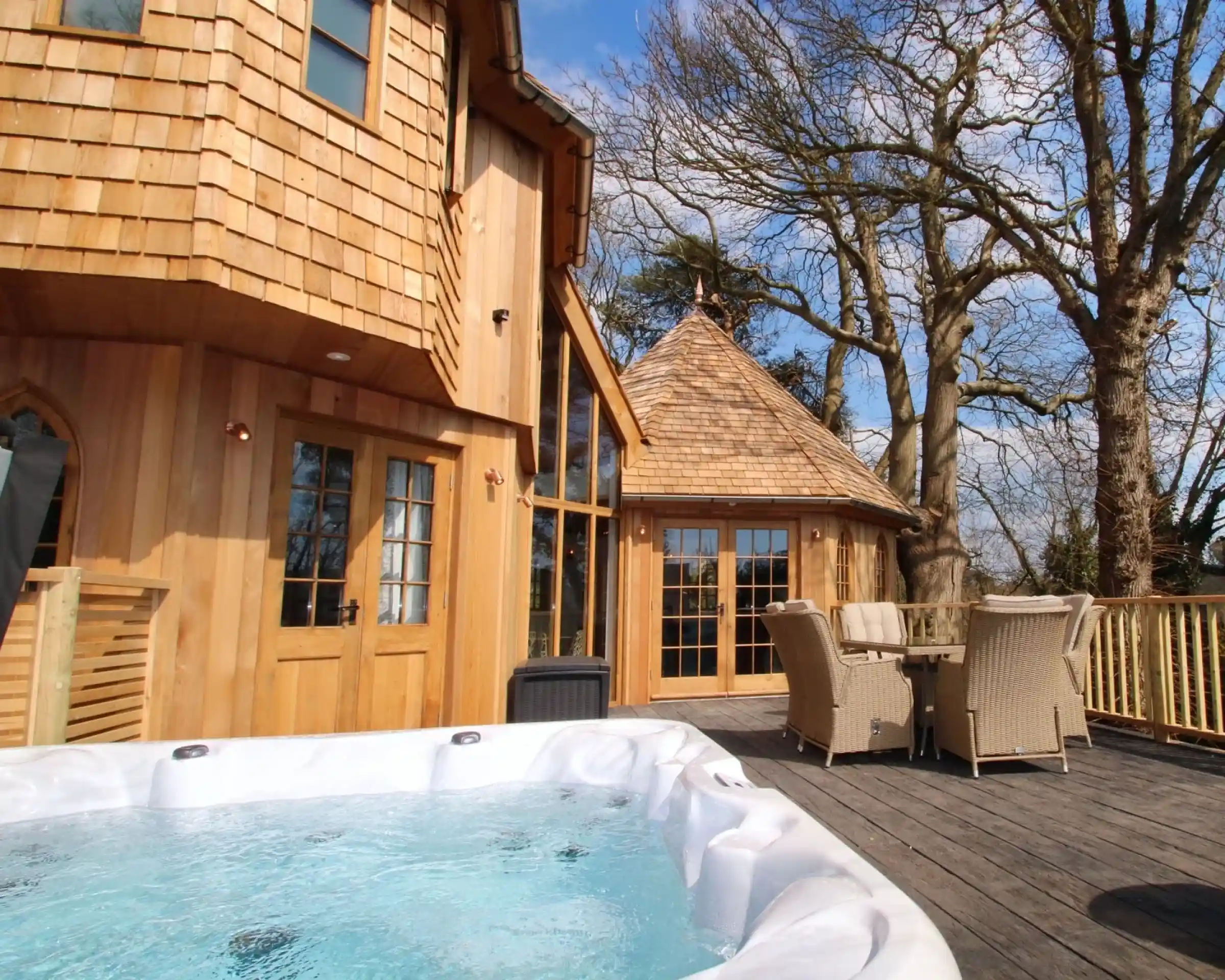 A wooden deck features a hot tub in the foreground, with a charming, rounded wooden building in the background surrounded by trees. Sunlight illuminates the scene under a clear blue sky.
