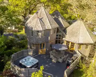 A charming wooden house with multiple conical roofs, surrounded by lush greenery. A spacious deck features a hot tub and outdoor dining area.