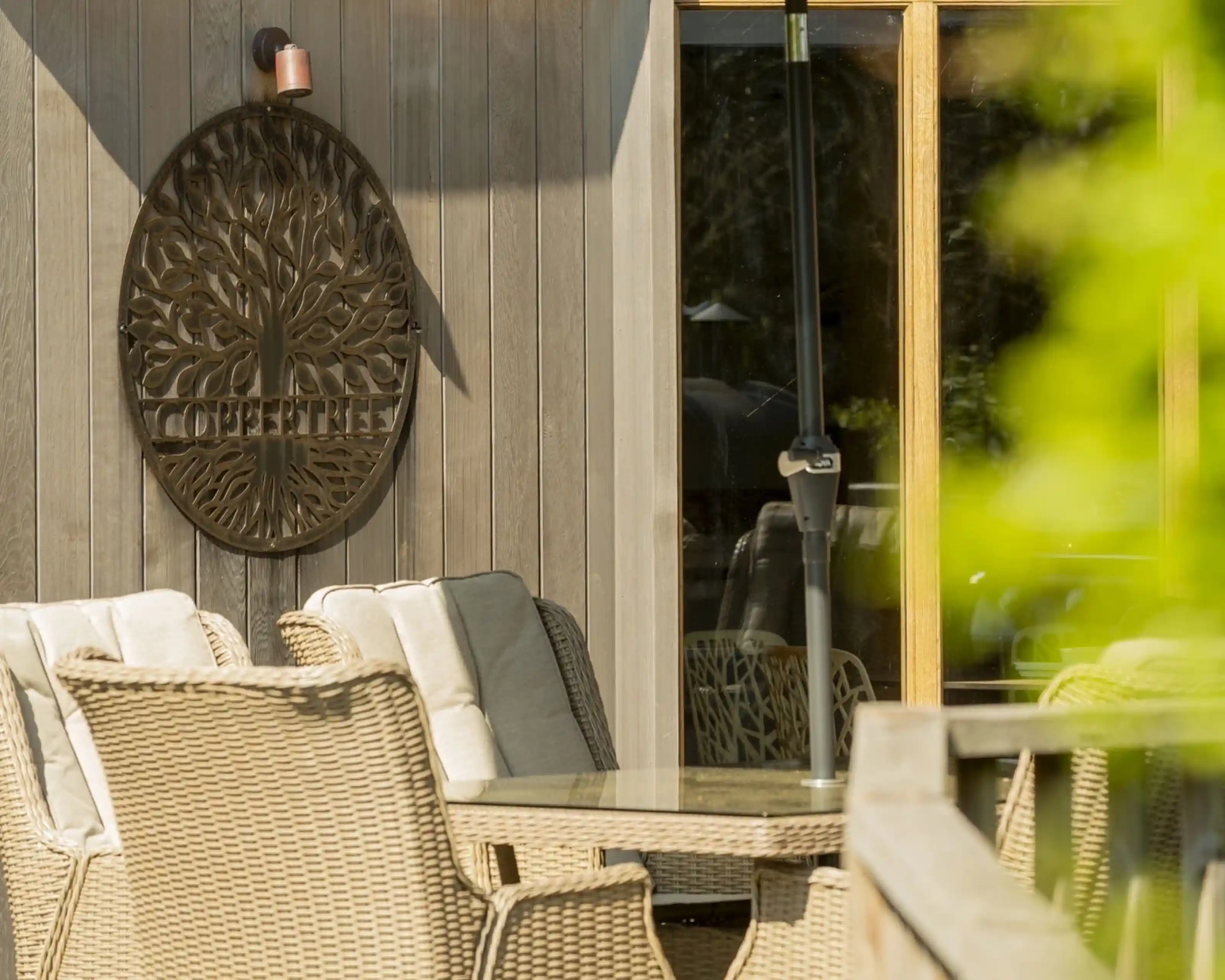 A cozy outdoor seating area featuring wicker furniture and a decorative tree wall art, with a large window reflecting the surroundings.