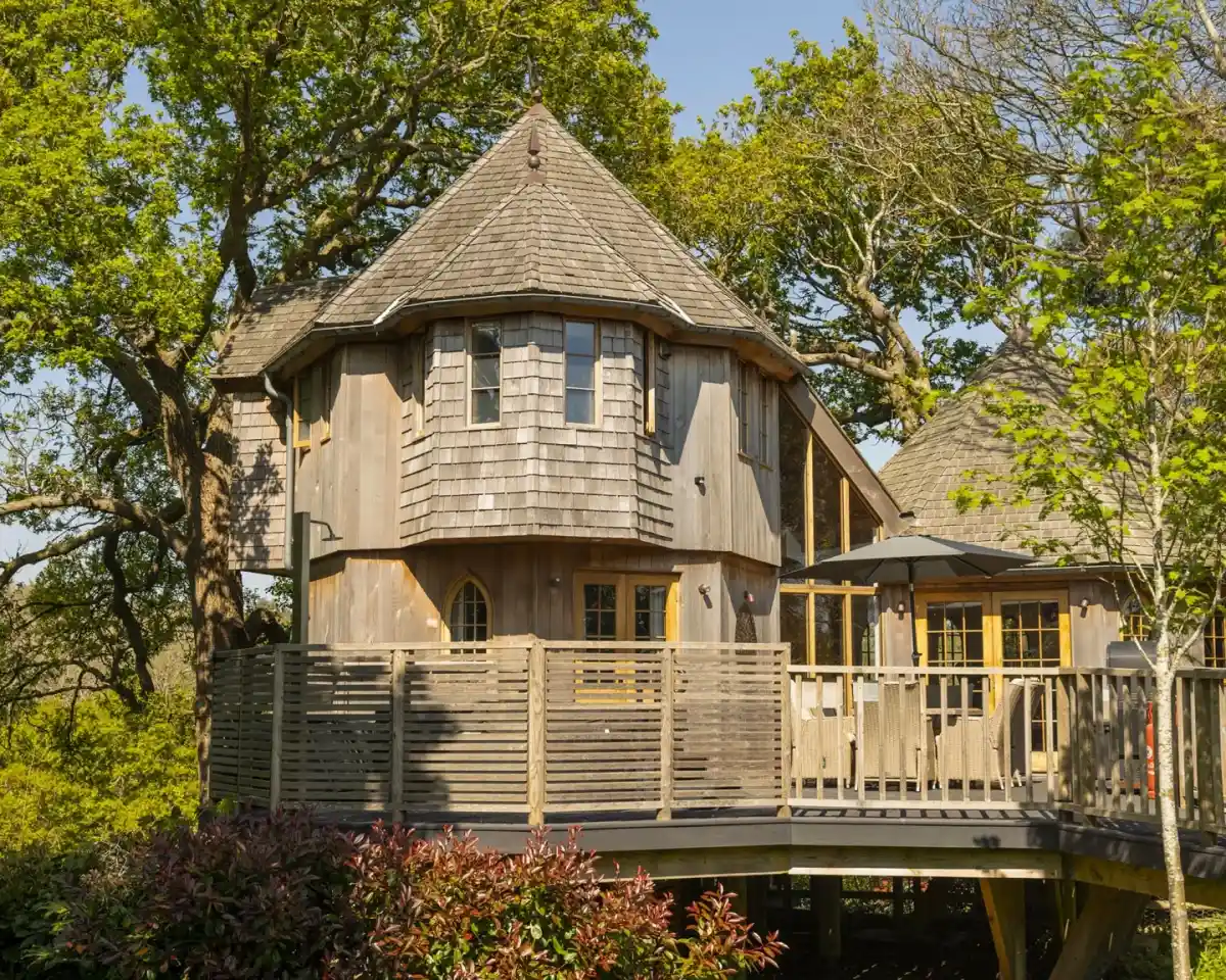 A unique octagonal wooden house surrounded by trees, featuring a wraparound deck and large windows. The exterior has a shingled roof and light-colored wood siding. Green foliage and a clear blue sky add to the serene atmosphere.