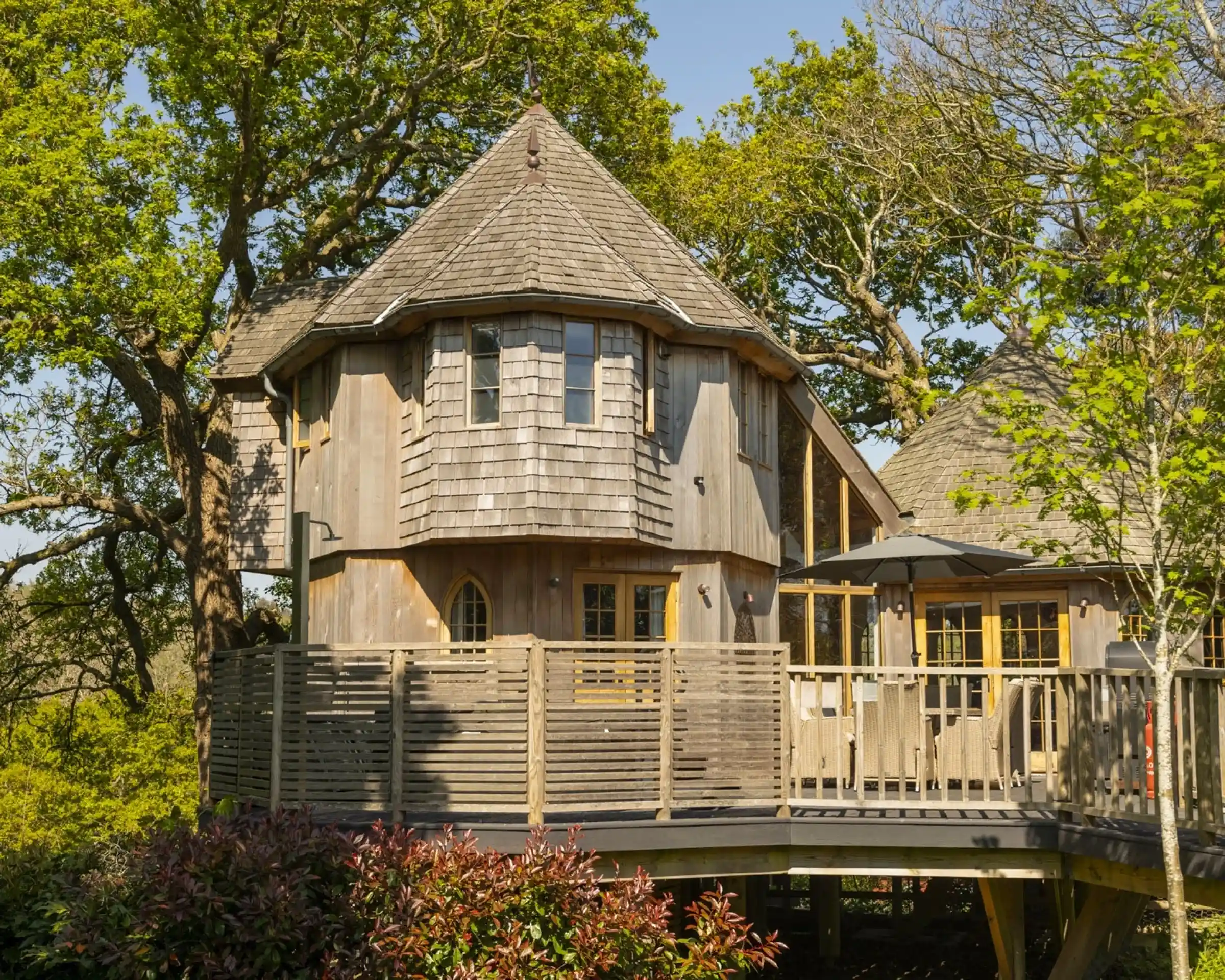 A unique octagonal wooden house surrounded by trees, featuring a wraparound deck and large windows. The exterior has a shingled roof and light-colored wood siding. Green foliage and a clear blue sky add to the serene atmosphere.