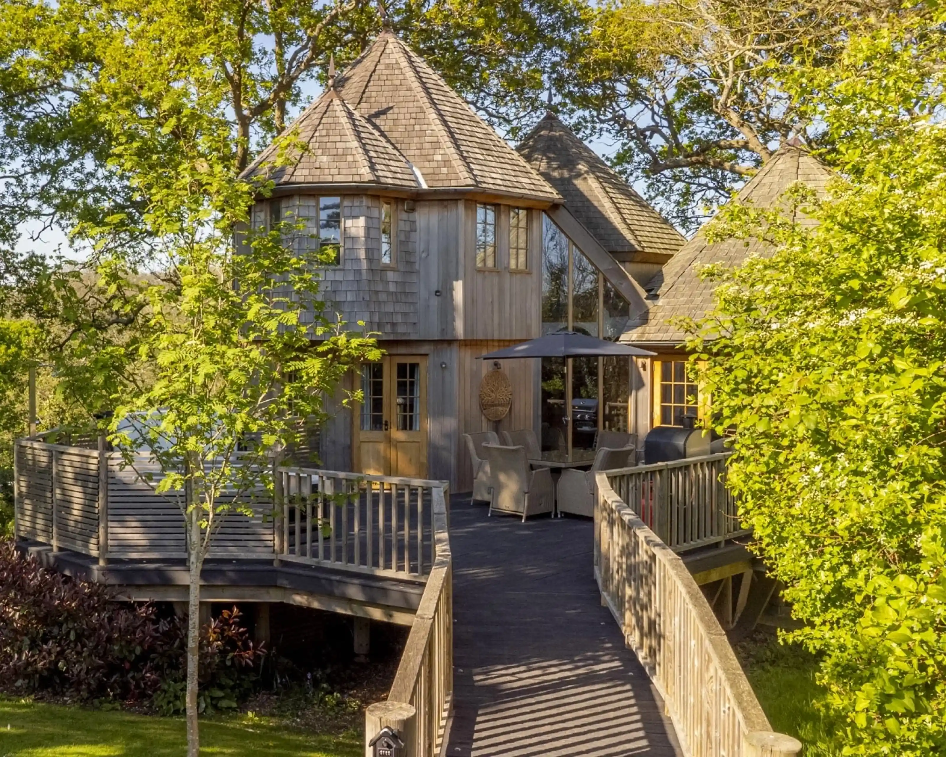 A charming wooden house with a unique turret, surrounded by lush greenery. A wooden deck leads to the entrance, featuring outdoor seating under a patio umbrella. Sunlight filters through the trees, creating a warm atmosphere.