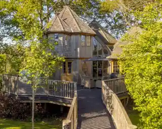 A charming wooden house with a unique turret, surrounded by lush greenery. A wooden deck leads to the entrance, featuring outdoor seating under a patio umbrella. Sunlight filters through the trees, creating a warm atmosphere.