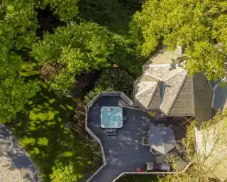 Aerial view of a house with a hot tub on a wooden deck surrounded by lush green trees. Sunlight casts shadows on the deck, enhancing the outdoor ambiance.