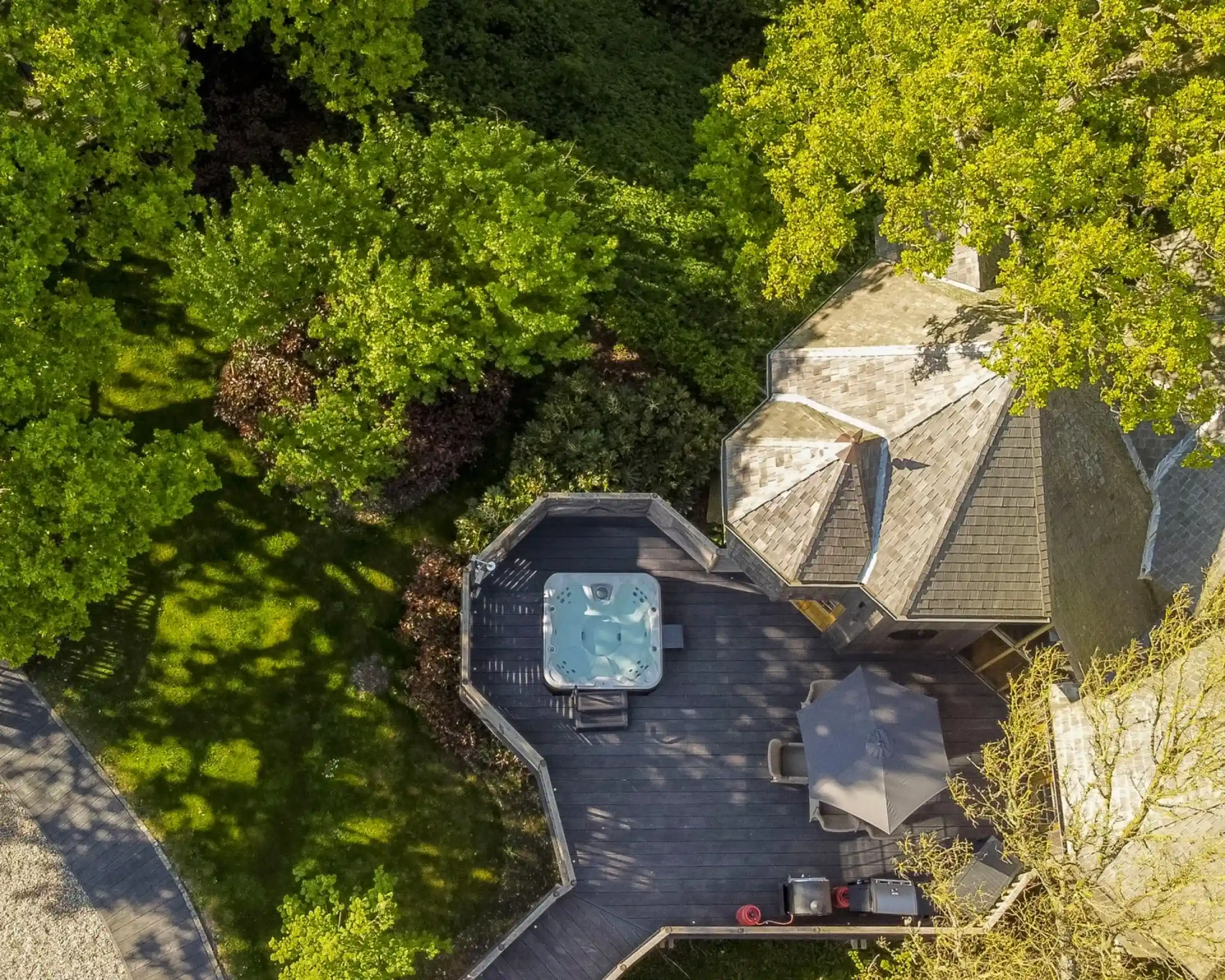 Aerial view of a house with a hot tub on a wooden deck surrounded by lush green trees. Sunlight casts shadows on the deck, enhancing the outdoor ambiance.