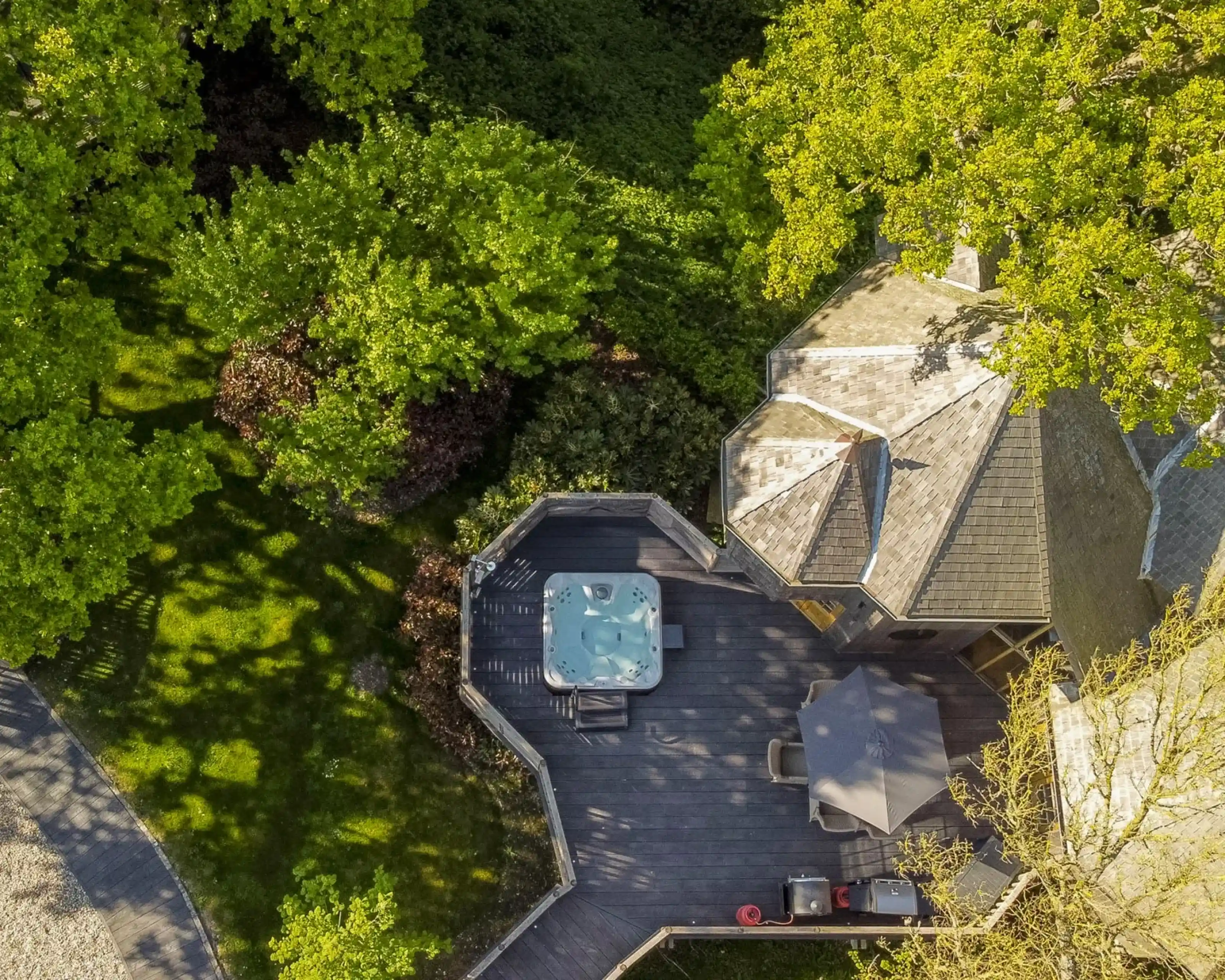 Aerial view of a house with a hot tub on a wooden deck surrounded by lush green trees. Sunlight casts shadows on the deck, enhancing the outdoor ambiance.