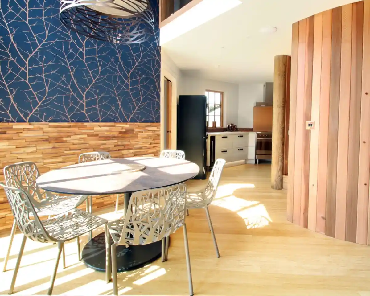 A modern dining area featuring a round table with intricate metal chairs, positioned next to a wall adorned with a tree branch pattern. Natural light streams in through large windows, highlighting wooden accents in the space and a contemporary kitchen in the background.