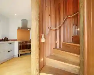 Wooden staircase with a rope handrail, leading to an upper level, alongside a modern kitchen featuring stainless steel appliances and warm wooden cabinetry.