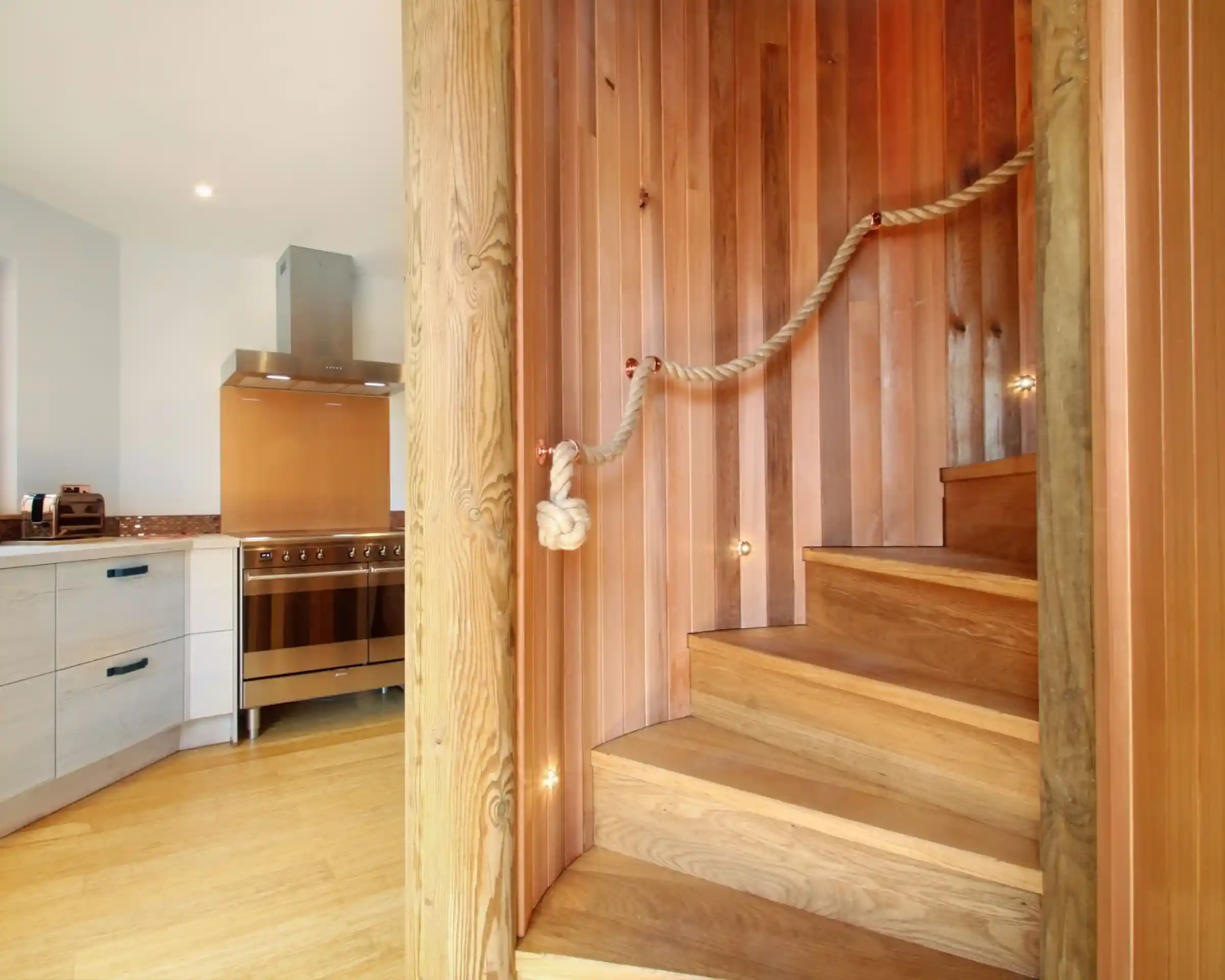 Wooden staircase with a rope handrail, leading to an upper level, alongside a modern kitchen featuring stainless steel appliances and warm wooden cabinetry.