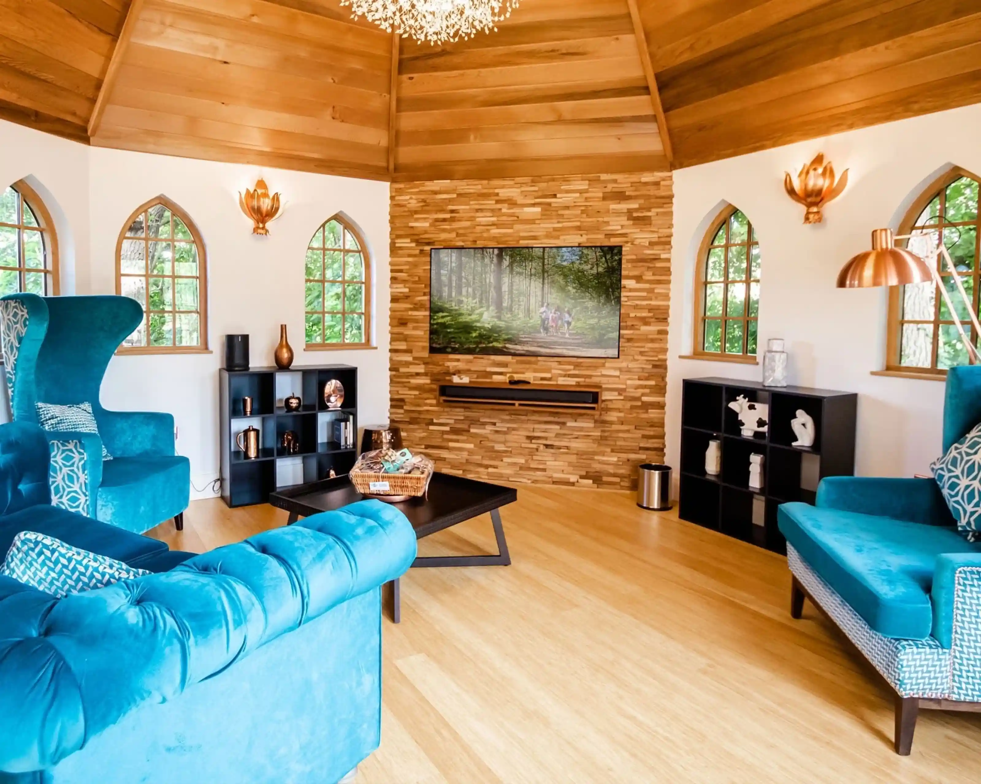 A cozy living room featuring blue velvet chairs and a black coffee table. A stone accent wall holds a flat-screen TV, while large arched windows provide natural light. Stylish decor includes modern lighting fixtures and decorative objects on shelves.