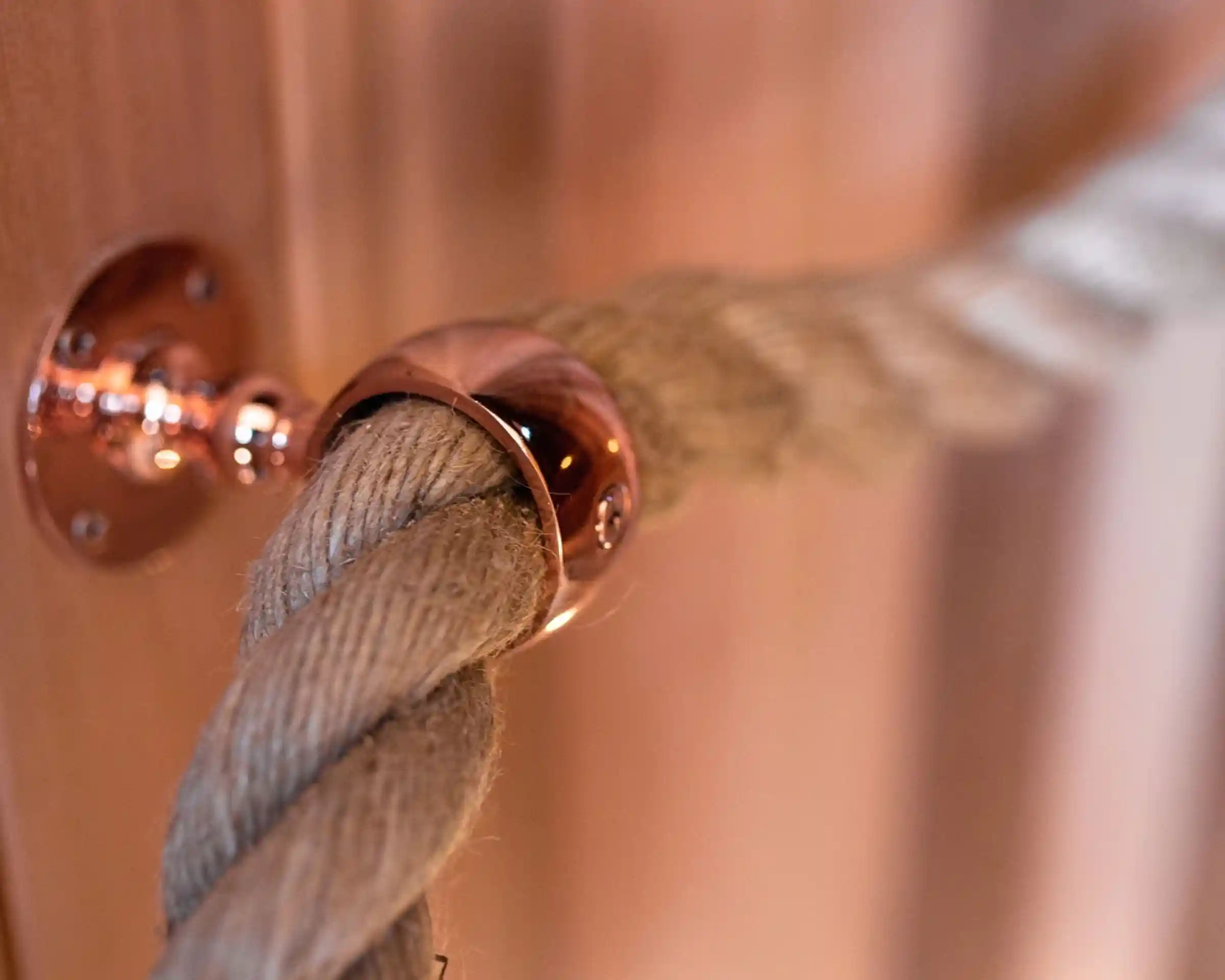 A thick natural rope secured with a shiny copper fixture attached to a wooden surface. The background has warm wood tones, creating a cozy atmosphere.