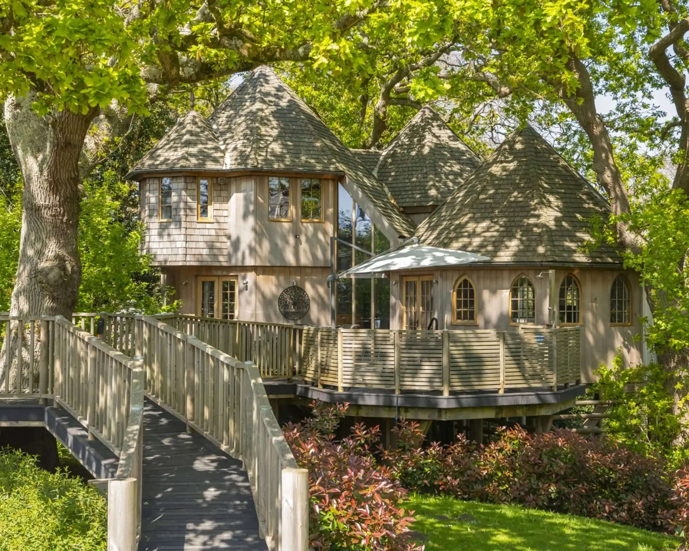 A charming wooden house with a unique, rounded architecture sits among lush greenery. It features a wraparound deck, large windows, and a sloping roof. A wooden walkway leads to the entrance, surrounded by vibrant plants and trees.