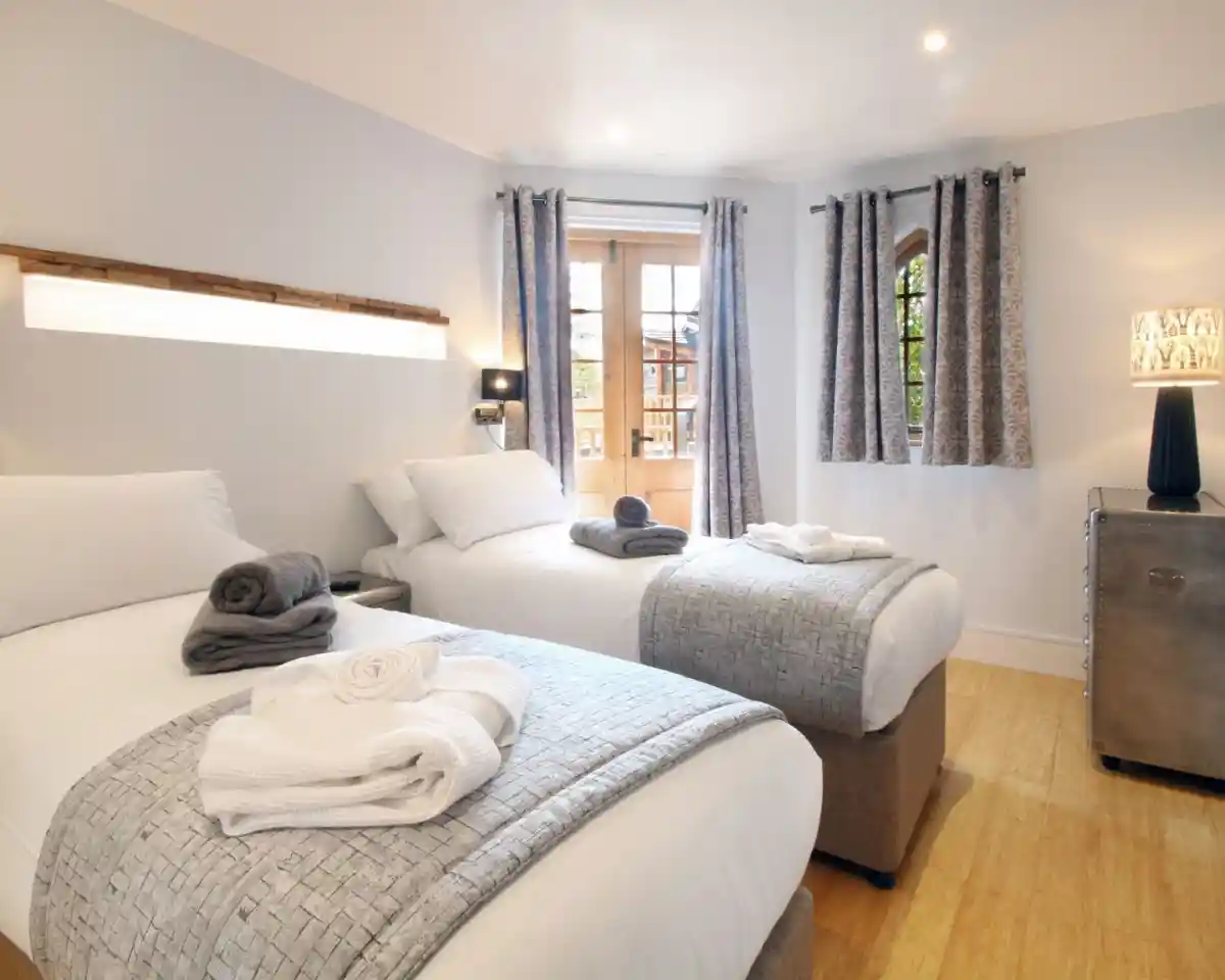 A bright hotel room featuring two twin beds with gray and white bedding. Soft towels are neatly arranged on the beds. Large windows with sheer curtains allow natural light to fill the space, and there is a stylish lamp on a small bedside table.