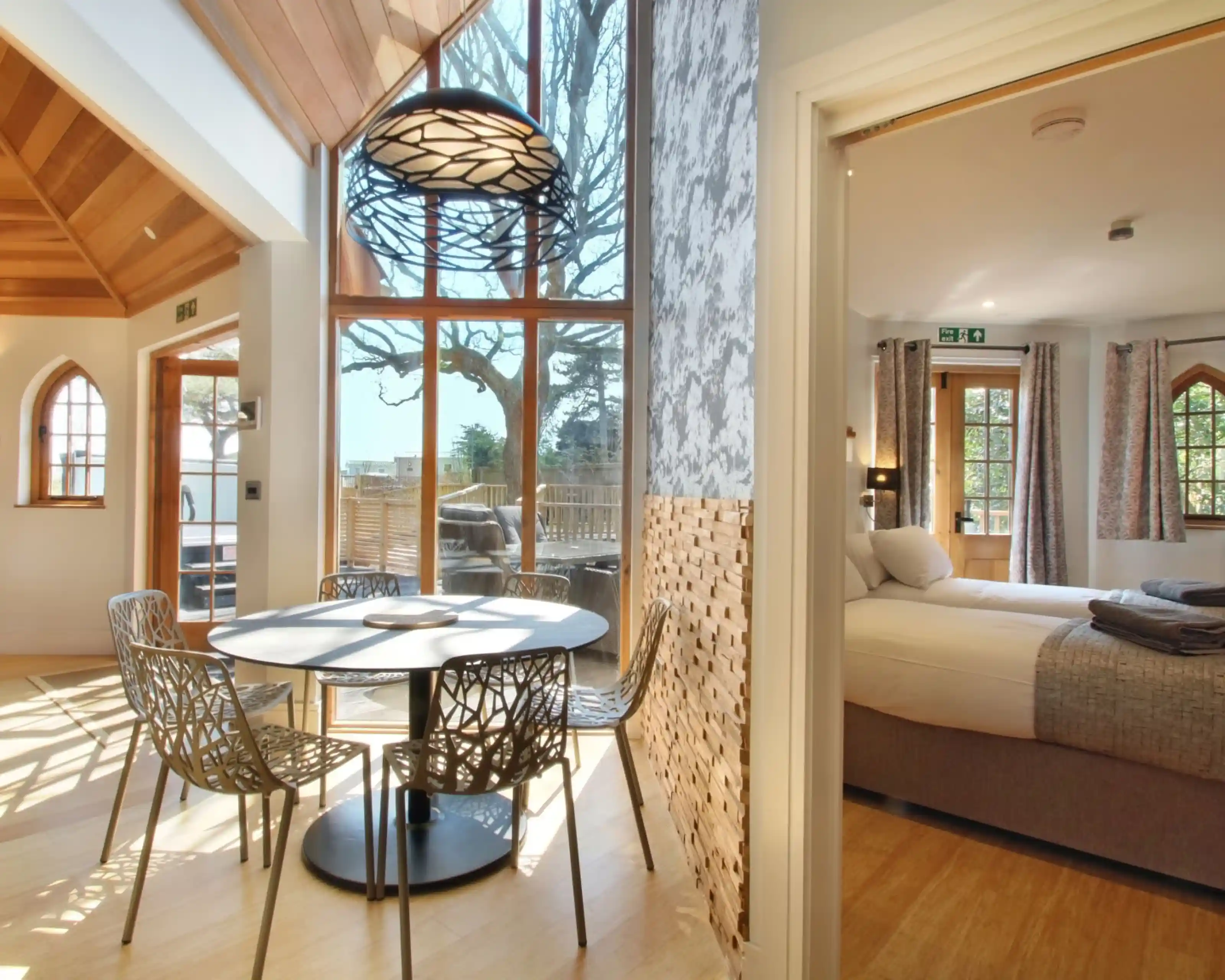 Bright and airy interior featuring a dining area with a circular table and modern chairs, large windows allowing natural light, and a view into a bedroom with a bed and soft decor.