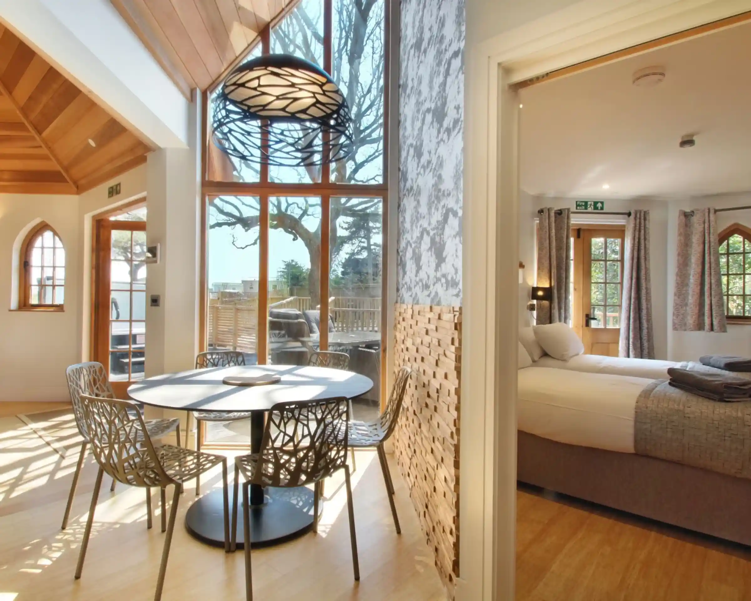 Bright and airy interior featuring a dining area with a circular table and modern chairs, large windows allowing natural light, and a view into a bedroom with a bed and soft decor.