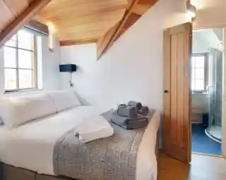 A cozy bedroom featuring a large bed with white linens and neatly arranged towels, a wooden ceiling with exposed beams, and a door leading to a bathroom with a glass shower.