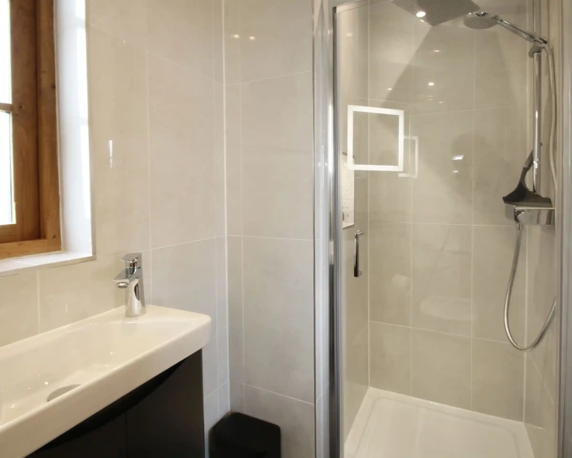 A modern bathroom featuring a glass shower enclosure, a sleek sink with a faucet, and a large window providing natural light. The walls are tiled with light-colored tiles, and there's a small black trash bin visible.