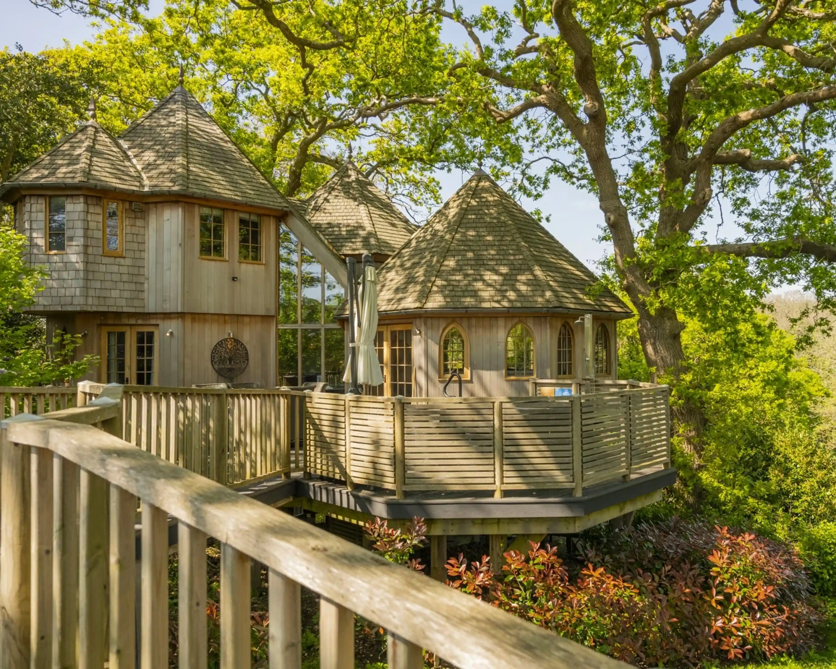 A unique wooden treehouse with a rounded design and multiple gabled roofs, surrounded by lush greenery. A wooden deck wraps around the structure, providing access to outdoor space. Sunlight filters through the trees, casting dappled shadows on the ground.