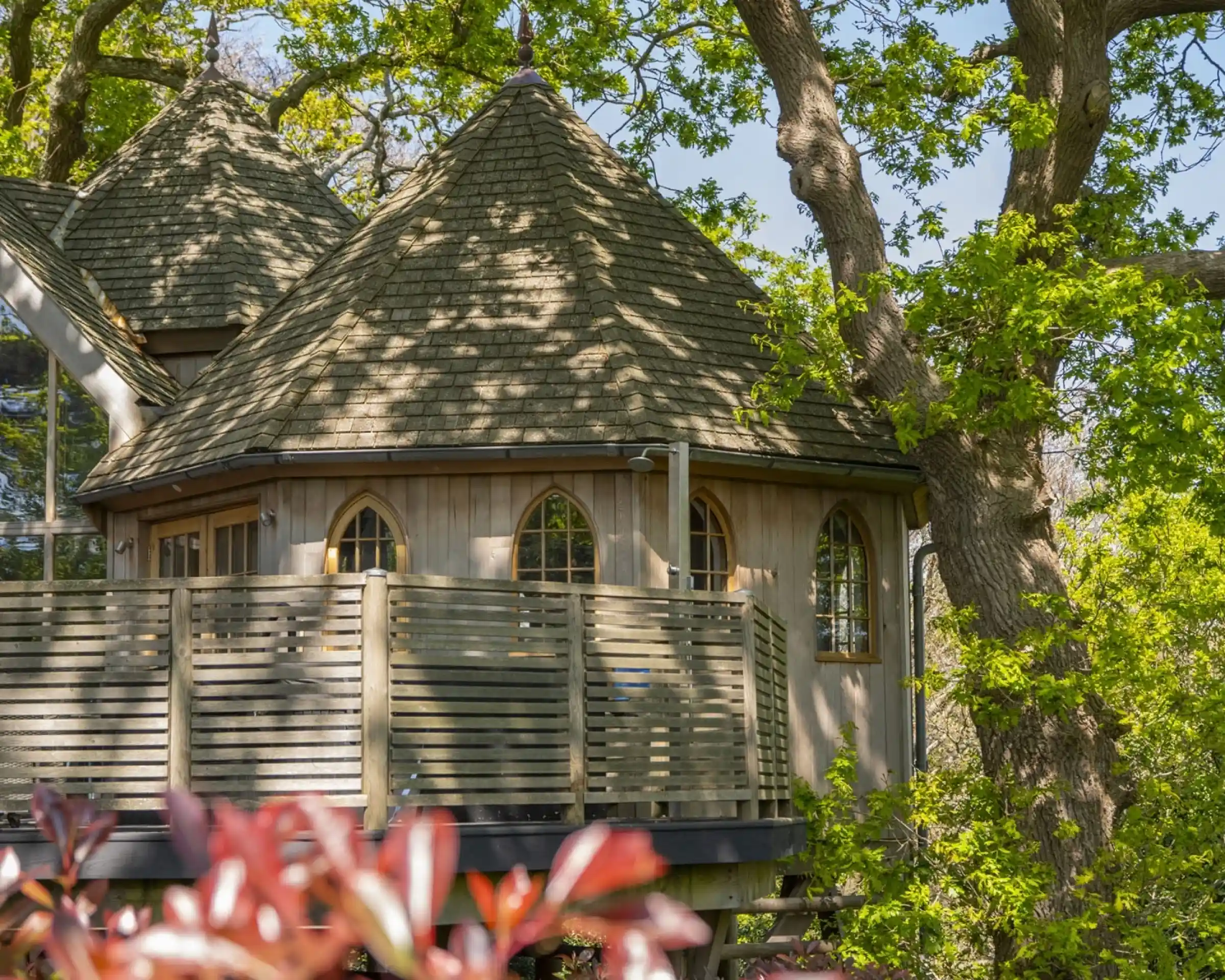 A charming treehouse with a shingled roof and wooden balcony nestled among vibrant green trees.