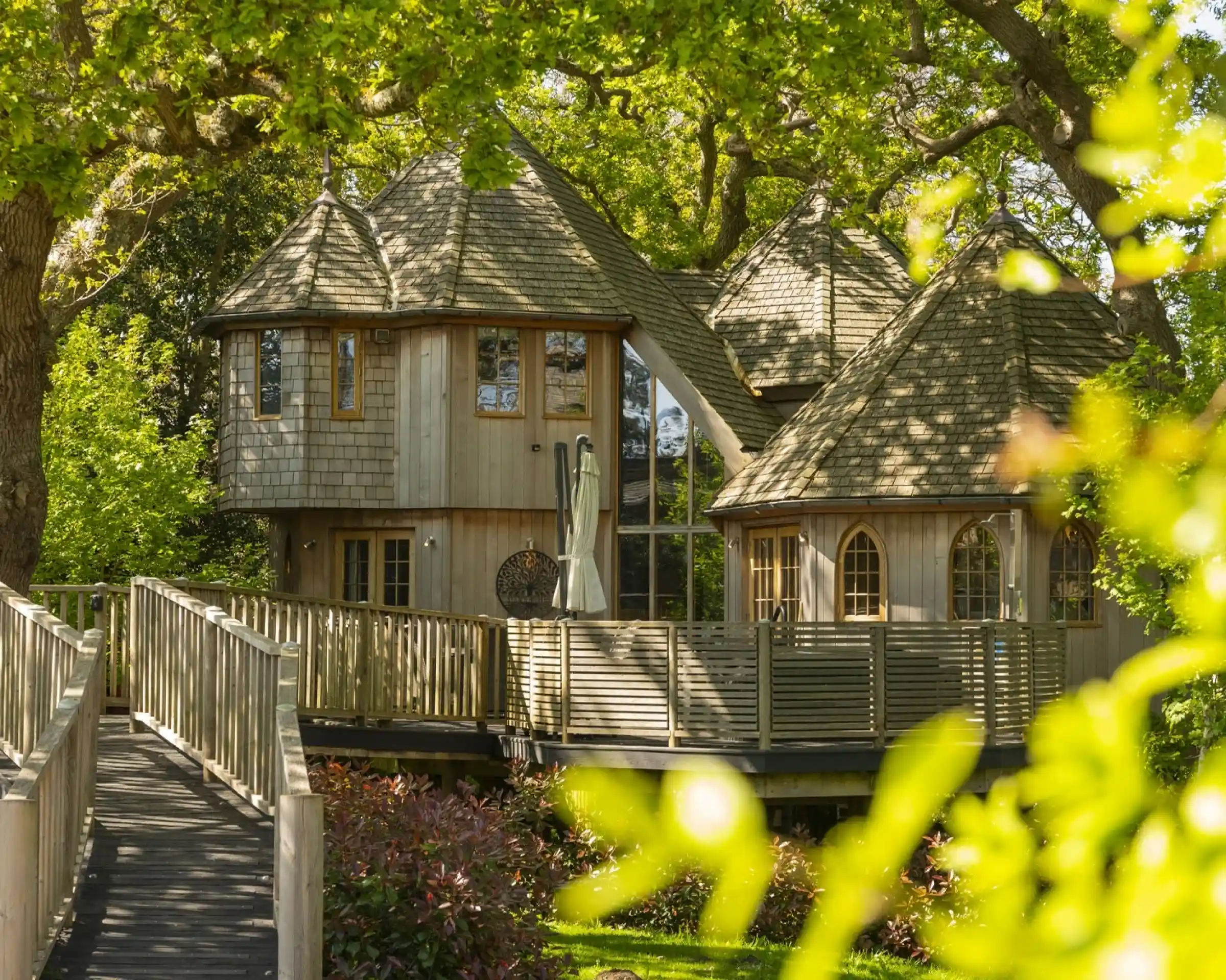 A unique wooden treehouse with multiple conical roofs surrounded by lush greenery. A wooden walkway leads up to the treehouse, which features large windows and a spacious deck.