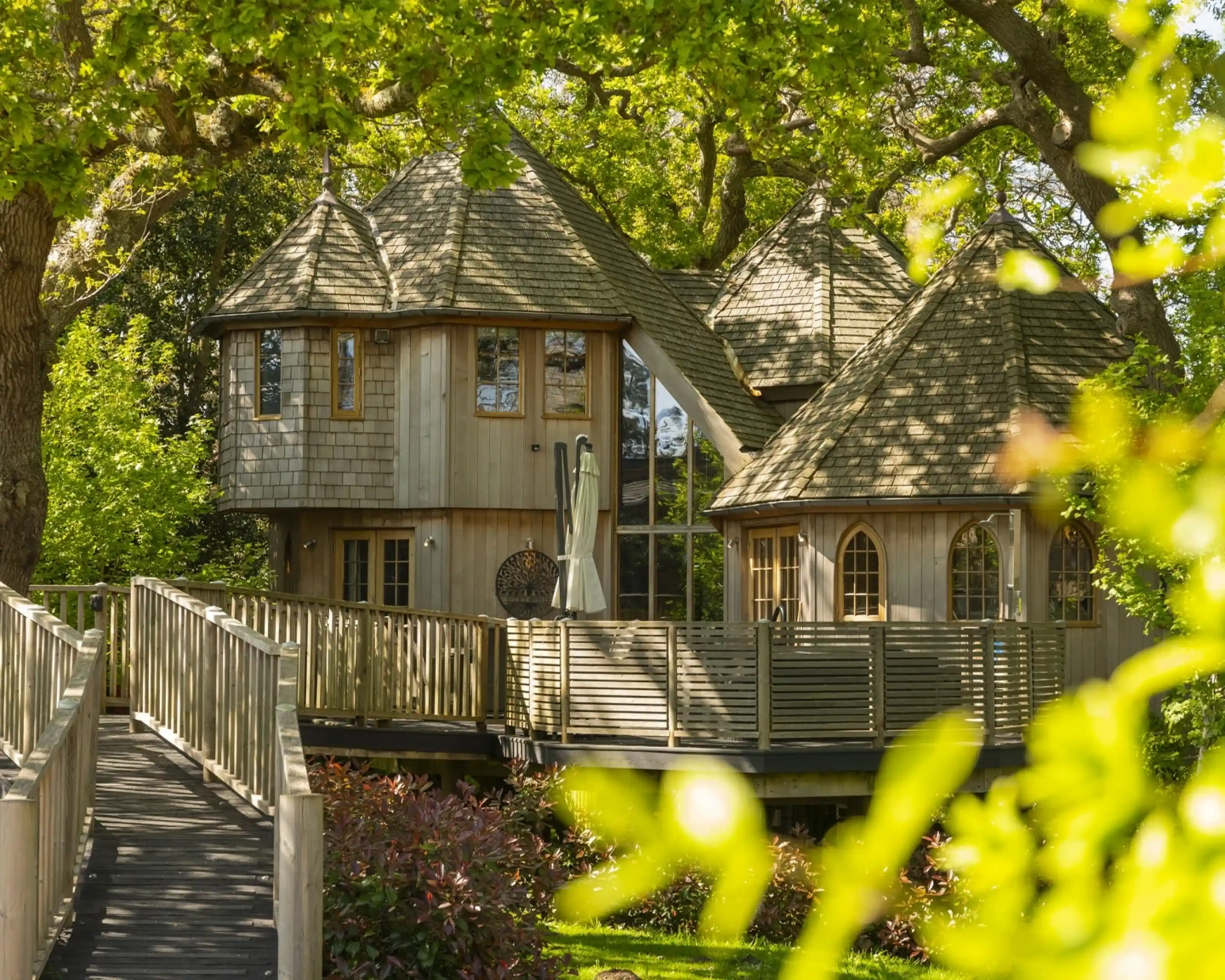 A unique wooden treehouse with multiple conical roofs surrounded by lush greenery. A wooden walkway leads up to the treehouse, which features large windows and a spacious deck.