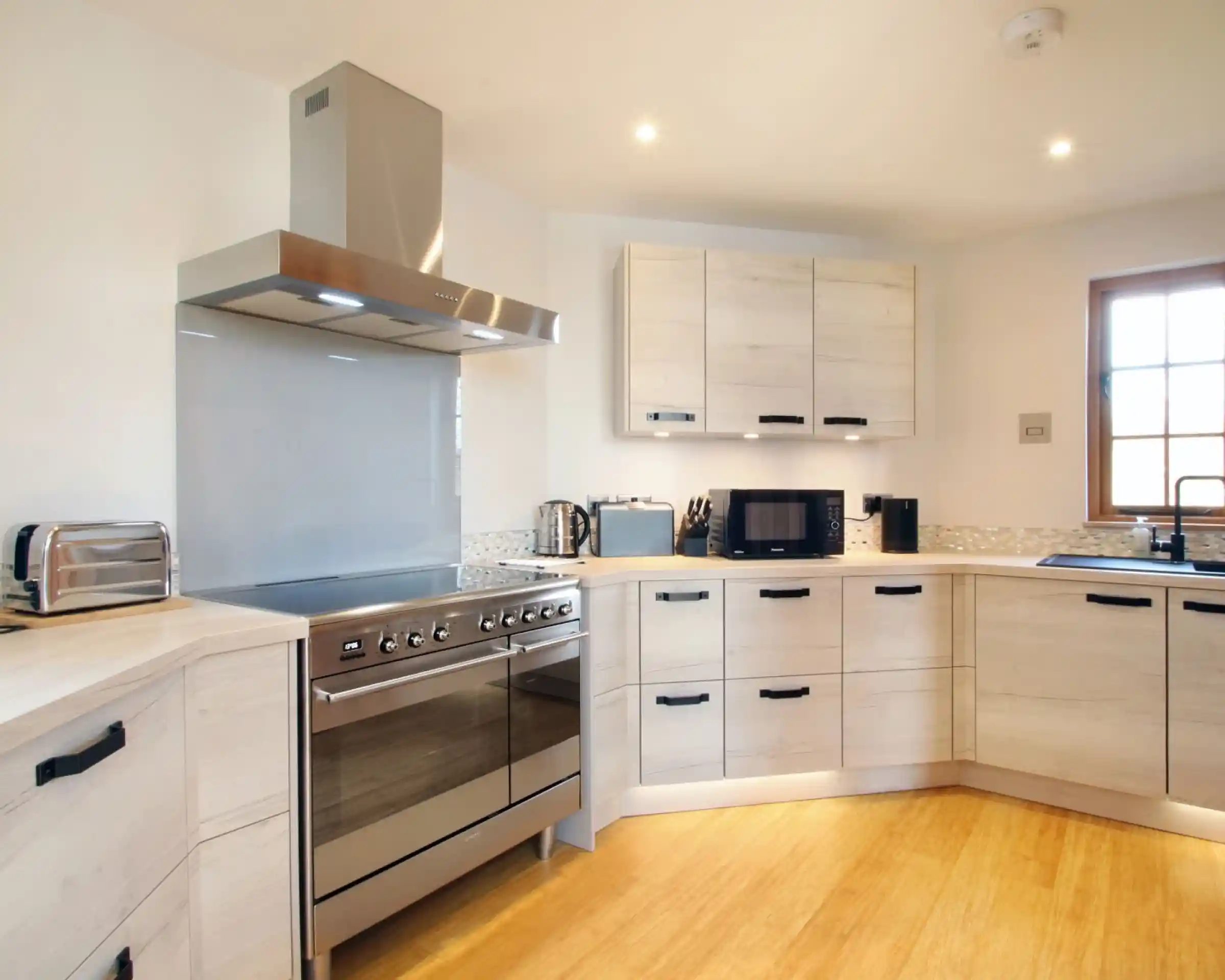 Modern kitchen featuring light wood cabinetry, stainless steel appliances, and a spacious countertop. A large oven range, hood, and various kitchen gadgets are visible. Natural light streams through a window.