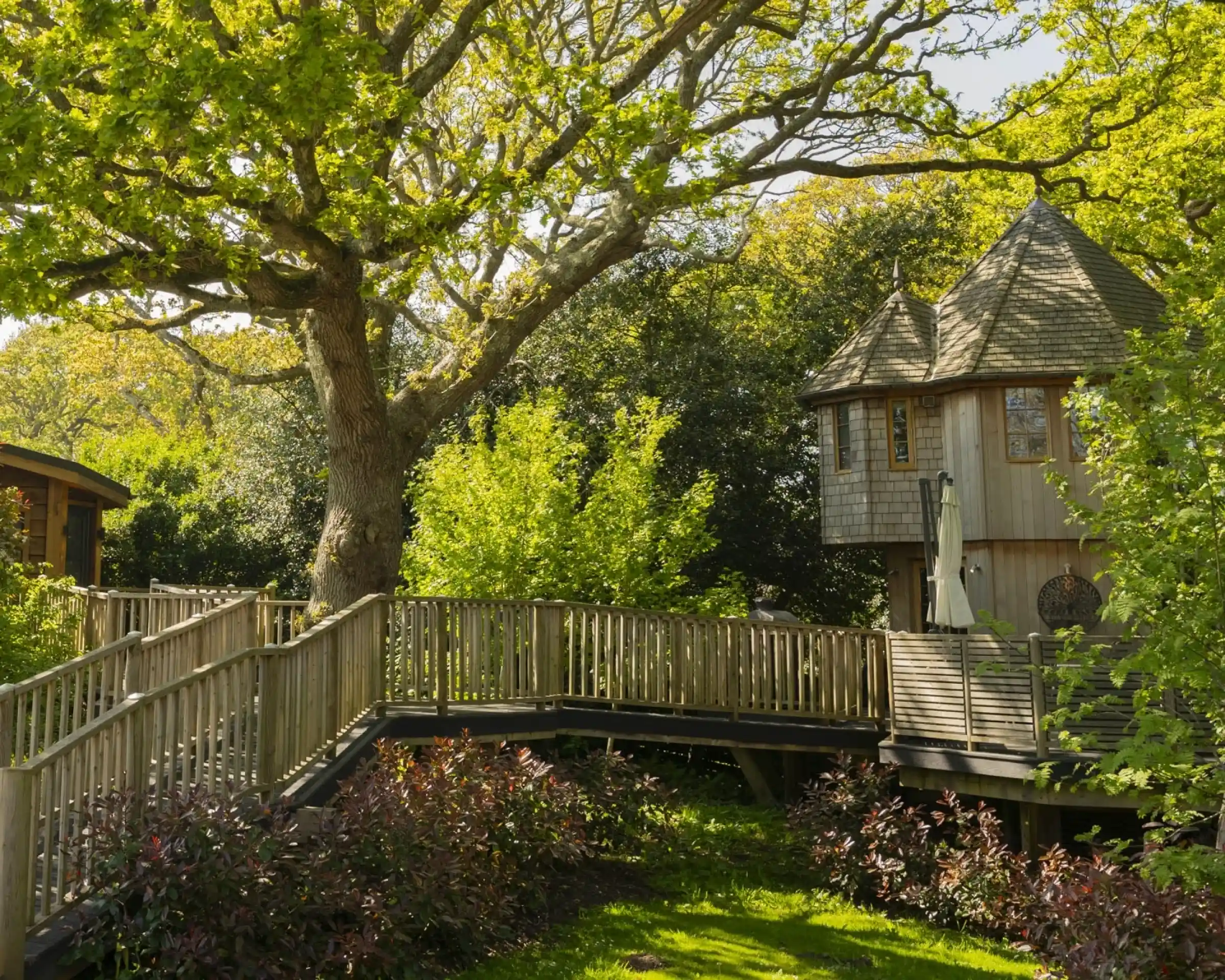 A wooden treehouse with a conical roof sits nestled among lush green trees and foliage, connected by a wooden walkway. Vibrant plants and grass surround the area, creating a serene outdoor environment.