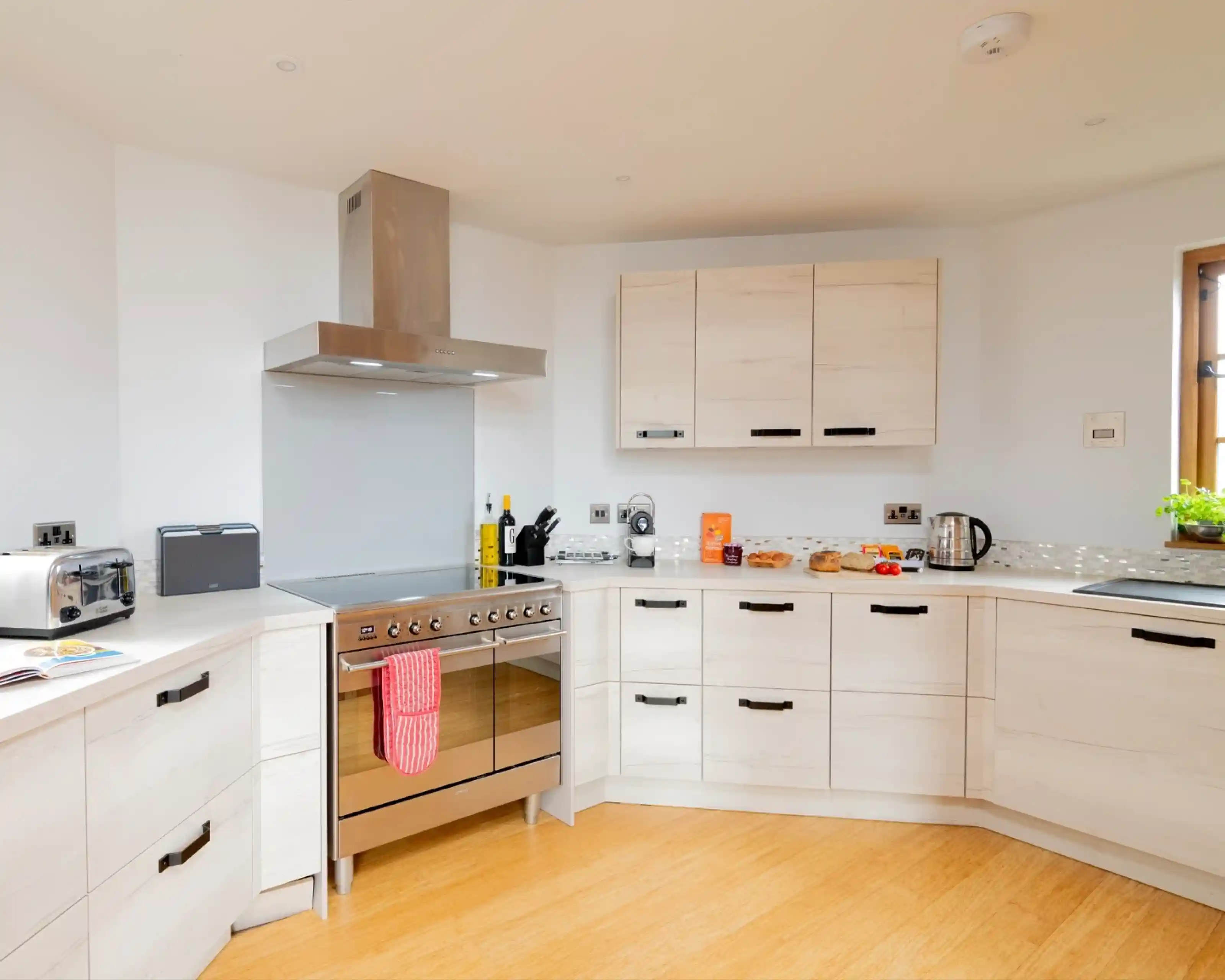 A modern kitchen with light wood cabinets, a stainless steel oven with a red towel hanging from it, and a variety of cooking utensils and appliances on the countertop. There is a window letting in natural light and a small table with snacks.