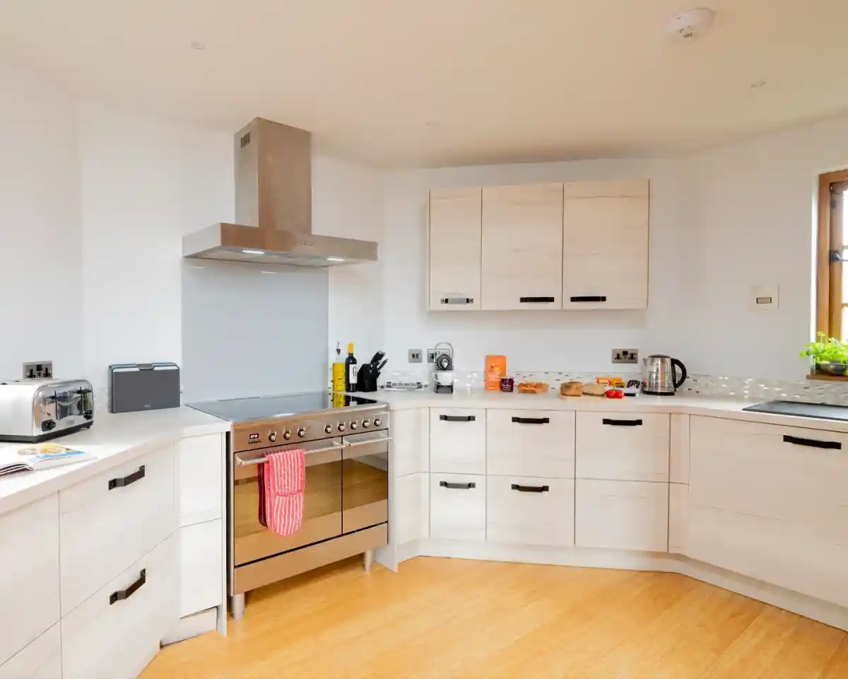 A modern kitchen with light wood cabinets, a stainless steel oven with a red towel hanging from it, and a variety of cooking utensils and appliances on the countertop. There is a window letting in natural light and a small table with snacks.