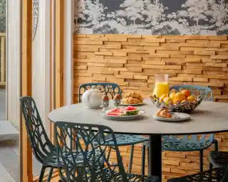 A dining table with a round concrete top surrounded by modern teal chairs. On the table are plates with fruit, croissants, and boiled eggs, next to a glass pitcher of orange juice. The backdrop features wooden paneling and a patterned wallpaper depicting trees.