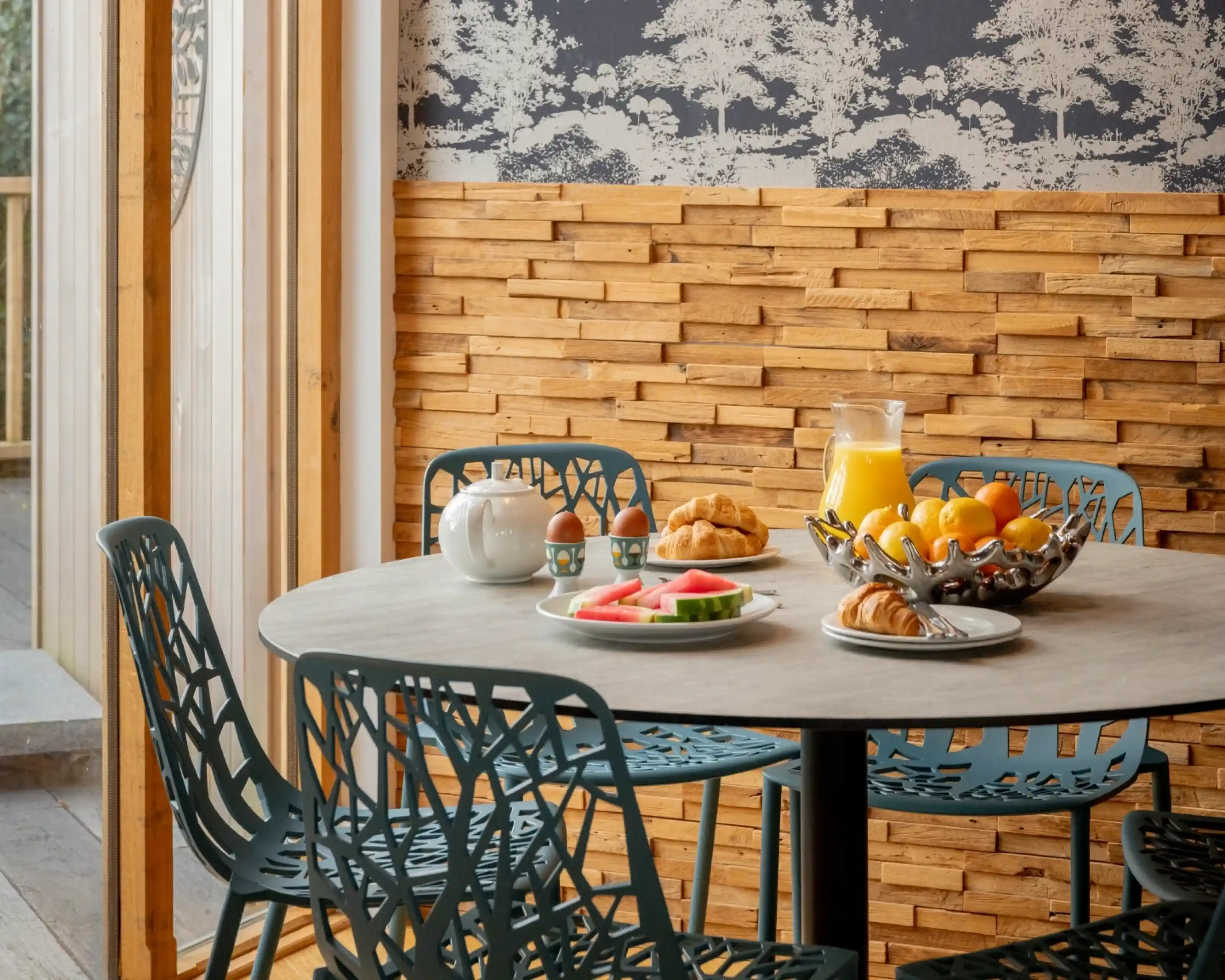 A dining table with a round concrete top surrounded by modern teal chairs. On the table are plates with fruit, croissants, and boiled eggs, next to a glass pitcher of orange juice. The backdrop features wooden paneling and a patterned wallpaper depicting trees.