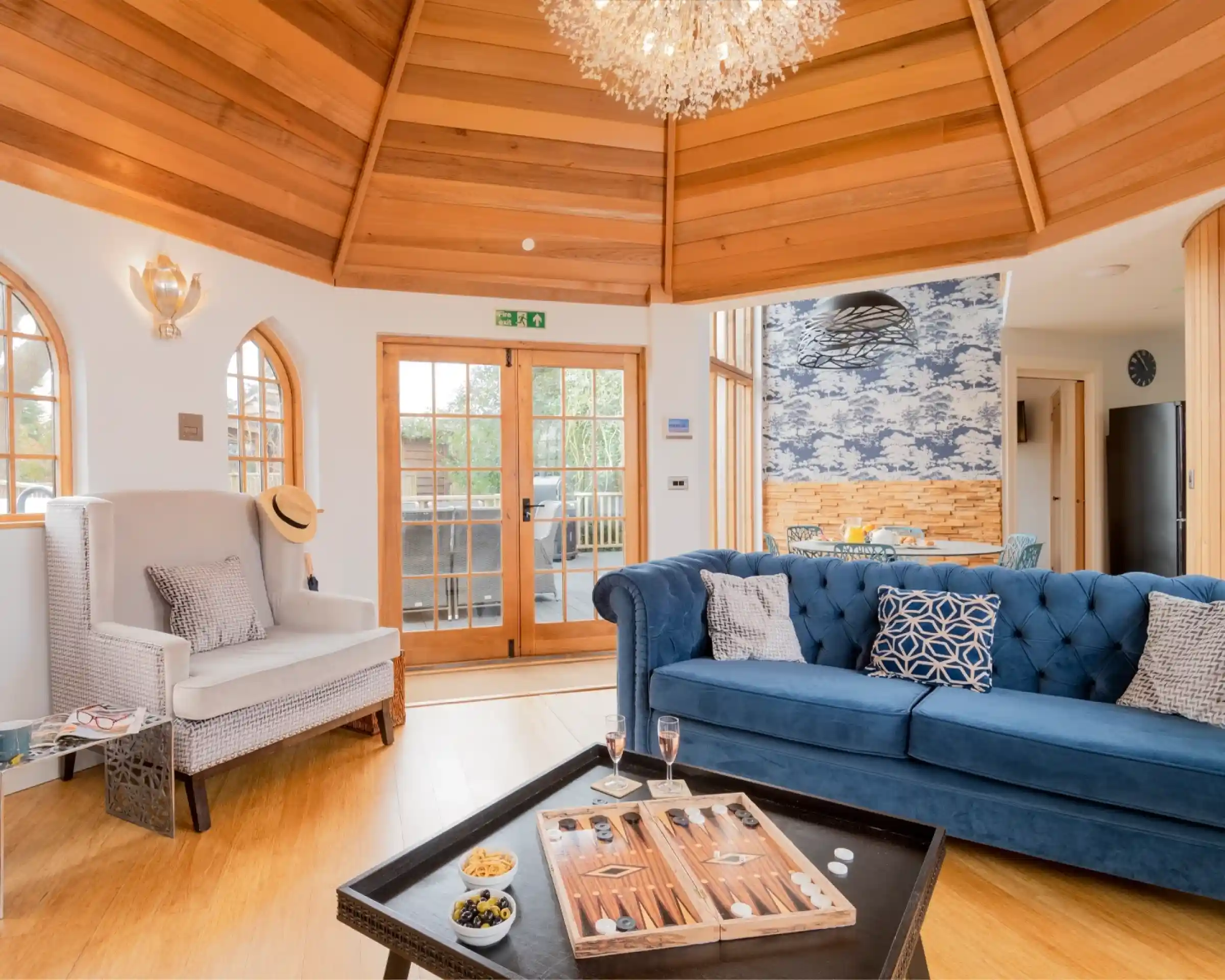 A cozy interior space featuring a wooden, vaulted ceiling. A blue velvet couch with decorative pillows faces a black coffee table with snacks and drinks. A light fixture hangs above, and there’s a stylish armchair to the left. Large windows provide natural light, and a doorway leads to an outdoor patio.
