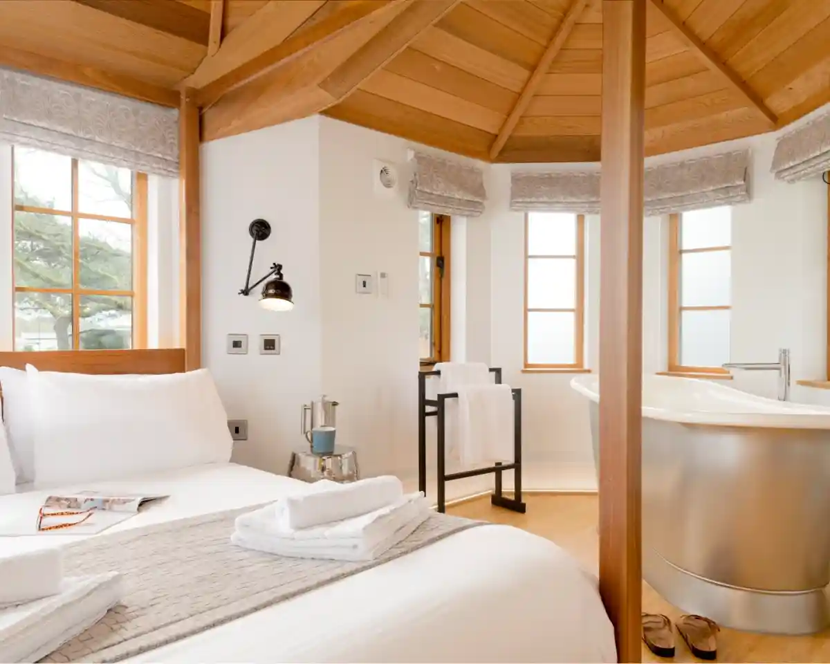 A cozy, circular room featuring a double bed with white linens, wooden accents, and a light wooden ceiling. Natural light streams in through multiple windows. A stylish bathtub is positioned near the windows, alongside a small table and two towels neatly arranged.